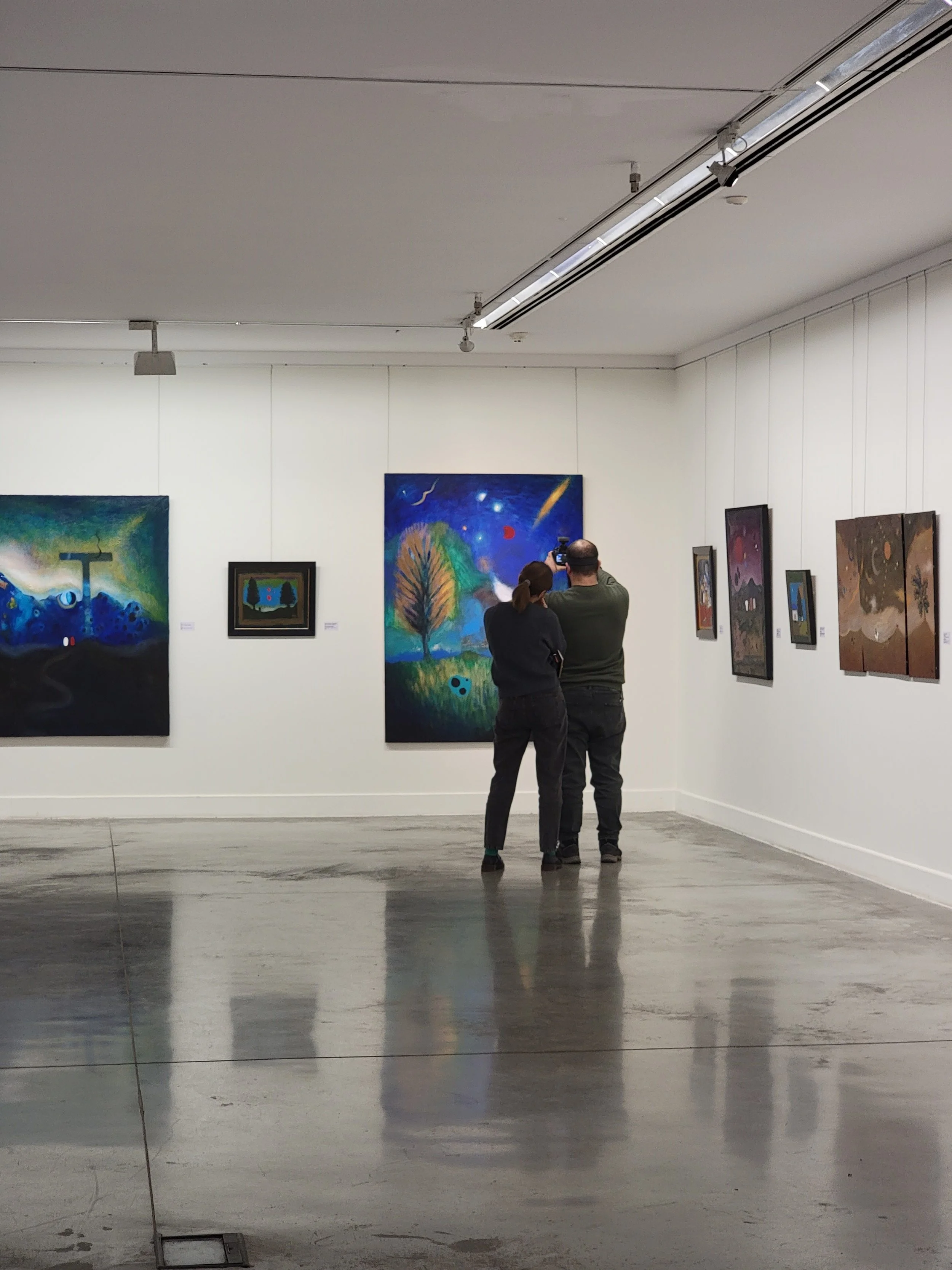 Two people are standing in front of colorful paintings in an art gallery, with one taking a photo.