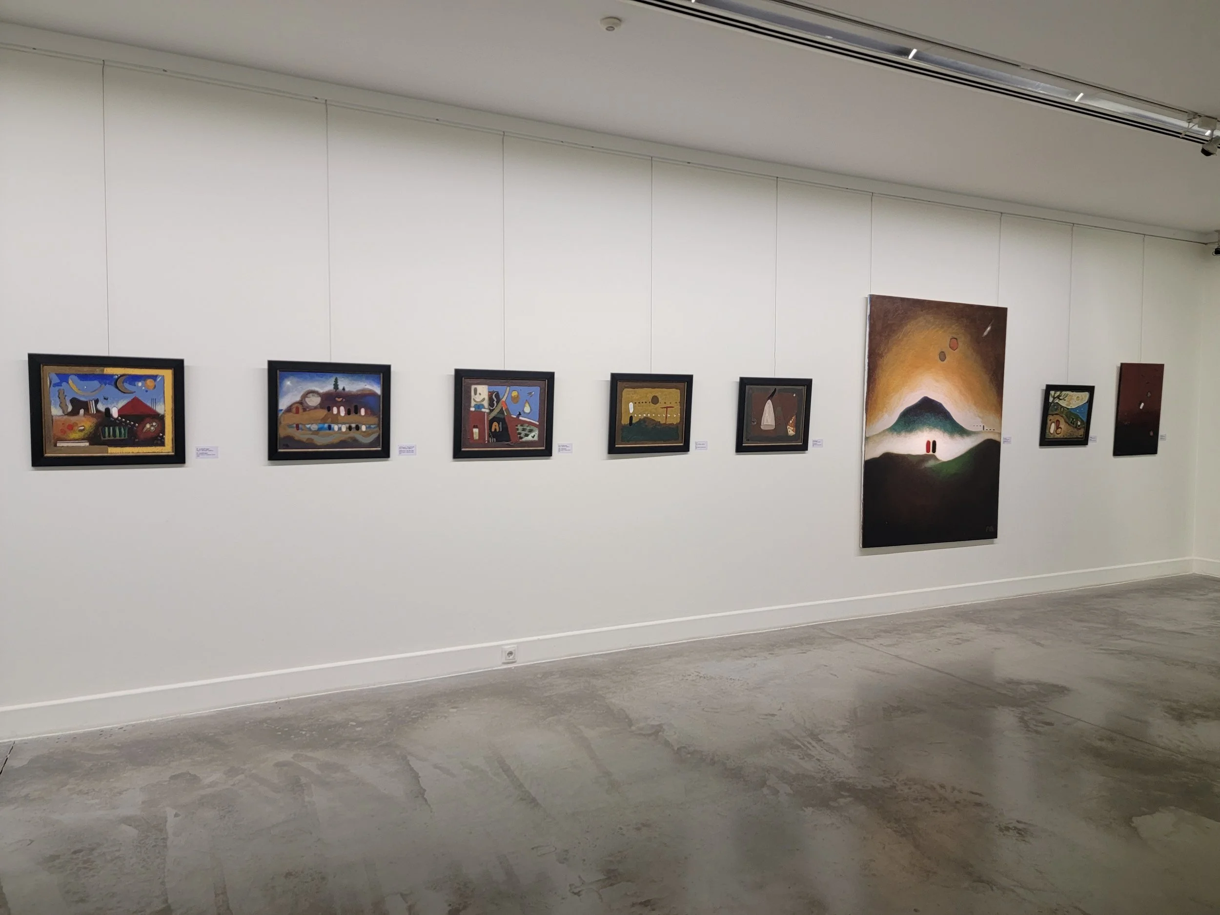 Art gallery with nine paintings displayed on a white wall, featuring abstract and surreal styles. The floor is concrete, and the ceiling has track lighting.