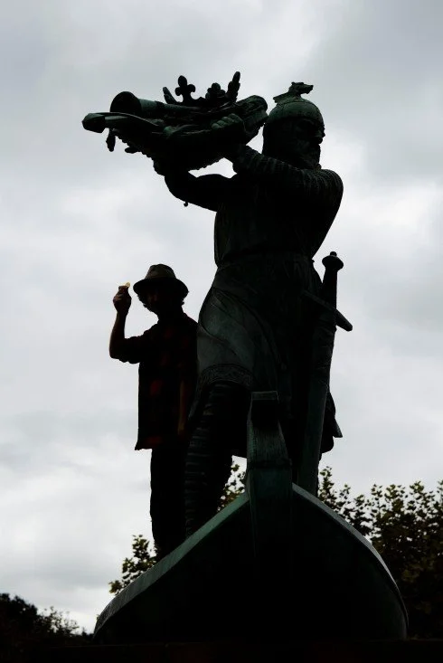 Silhouette of a person wearing a hat standing behind a large statue of a woman holding a ship modeled after the Statue of Liberty, against a cloudy sky.