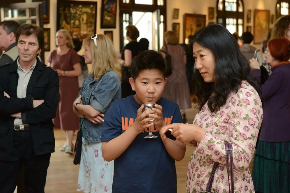 A group of people are at an art gallery , with paintings of Anton Balanchivadze. A woman and a boy in the foreground are looking at a device held by the woman. Other visitors are in the background, some watching and some engaging in conversation.