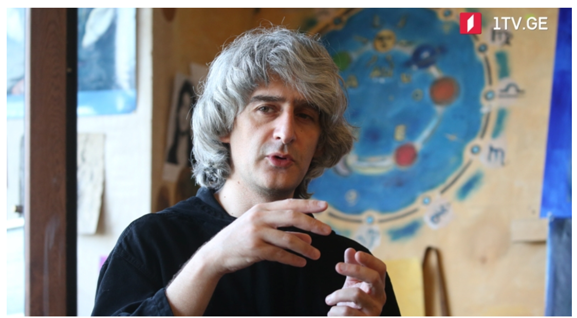 A man with gray, wavy hair speaking and gesturing with his hands in a room with a colorful cosmic-themed artwork on the wall.