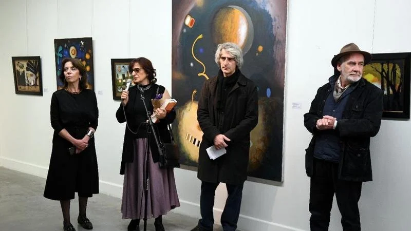 Four people standing in an art gallery, with various paintings on the white wall behind them. One woman is speaking into a microphone, while the others listen.