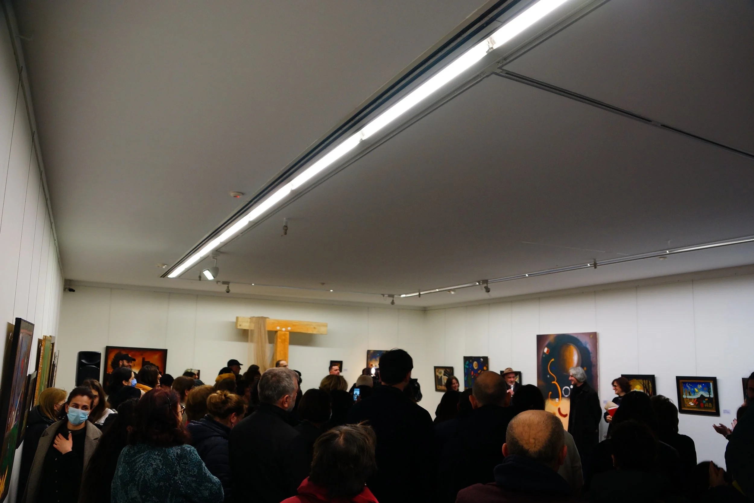 People gathered at an art gallery opening, looking at various paintings displayed on white walls, with some visitors wearing masks.