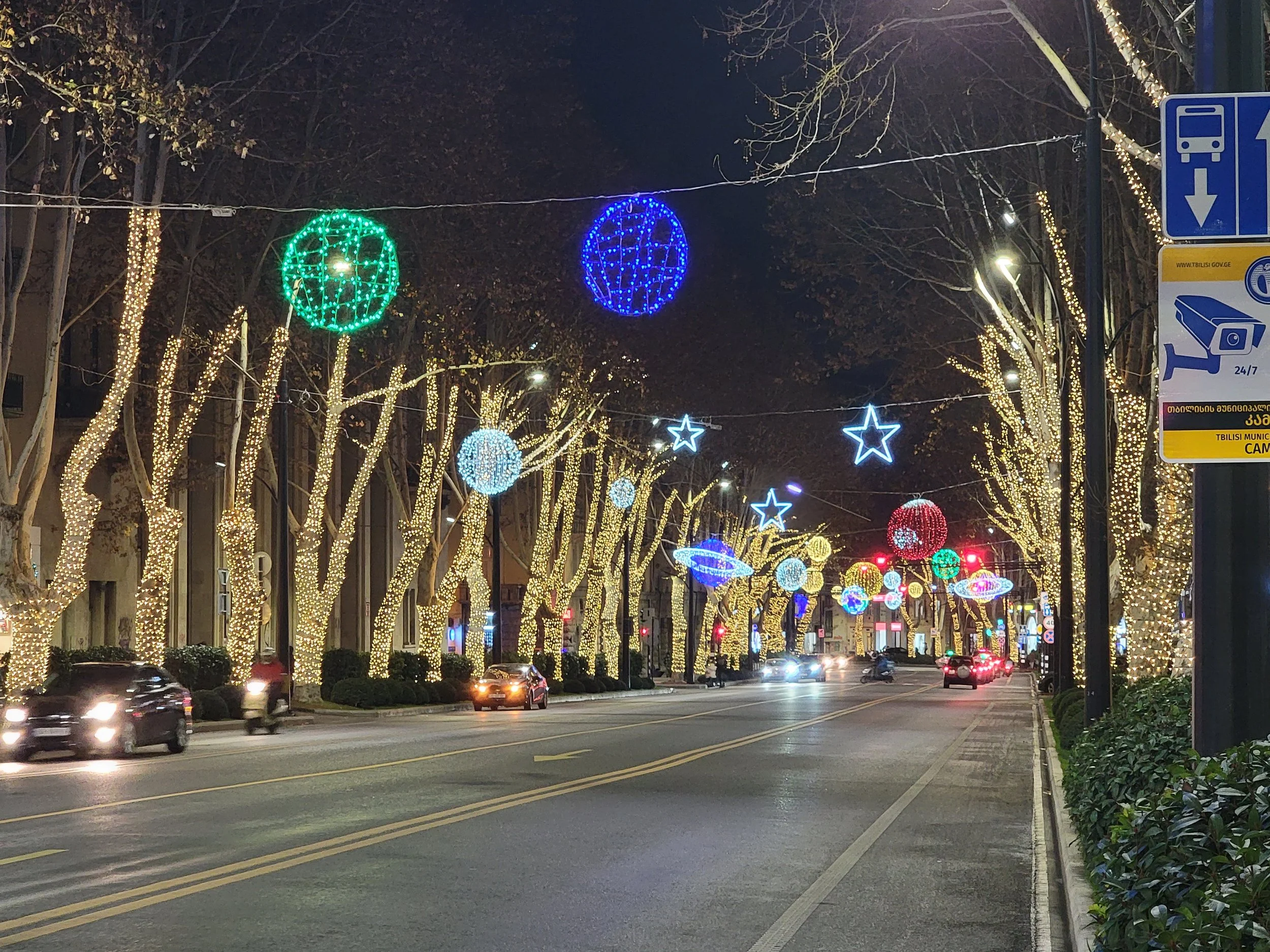 Street decorated with Christmas lights and ornaments at night, with cars and motorcycles on the road.