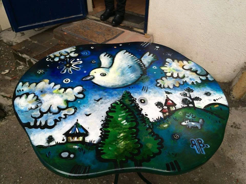 Colorful painted table featuring a white dove flying in a cloudy sky, surrounded by trees, houses, and a dog in a green landscape.