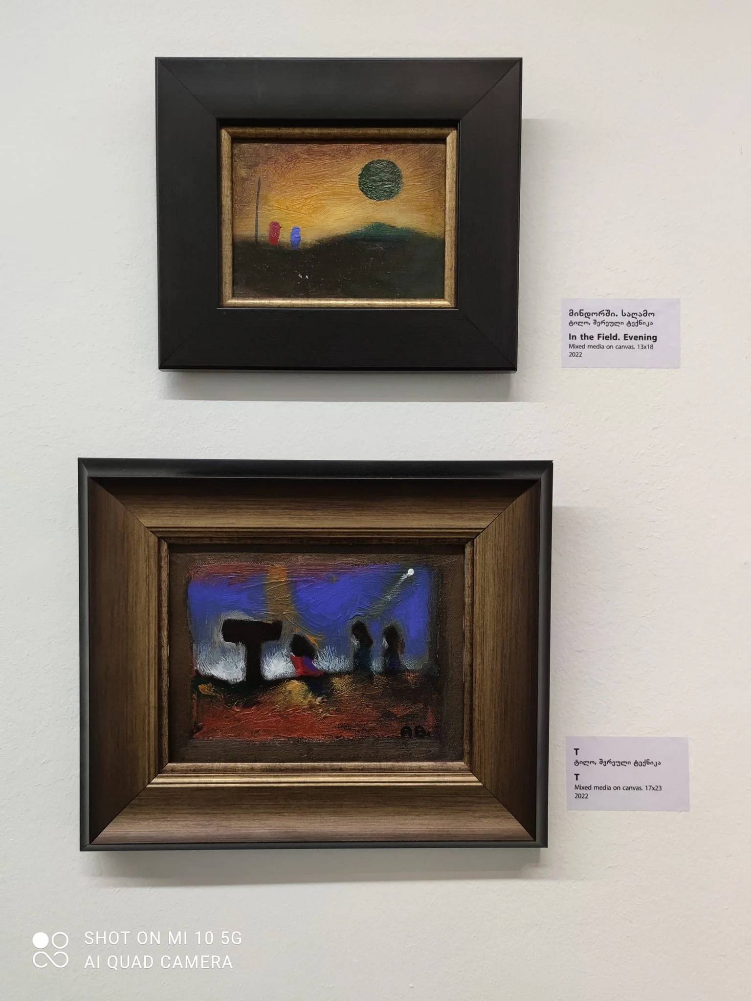 Two small framed paintings on a white wall. The upper painting depicts a sunset scene with a black sun, a small hill, and two colorful vertical objects, with a dark foreground. The lower painting shows an abstract scene with dark shapes against a blu