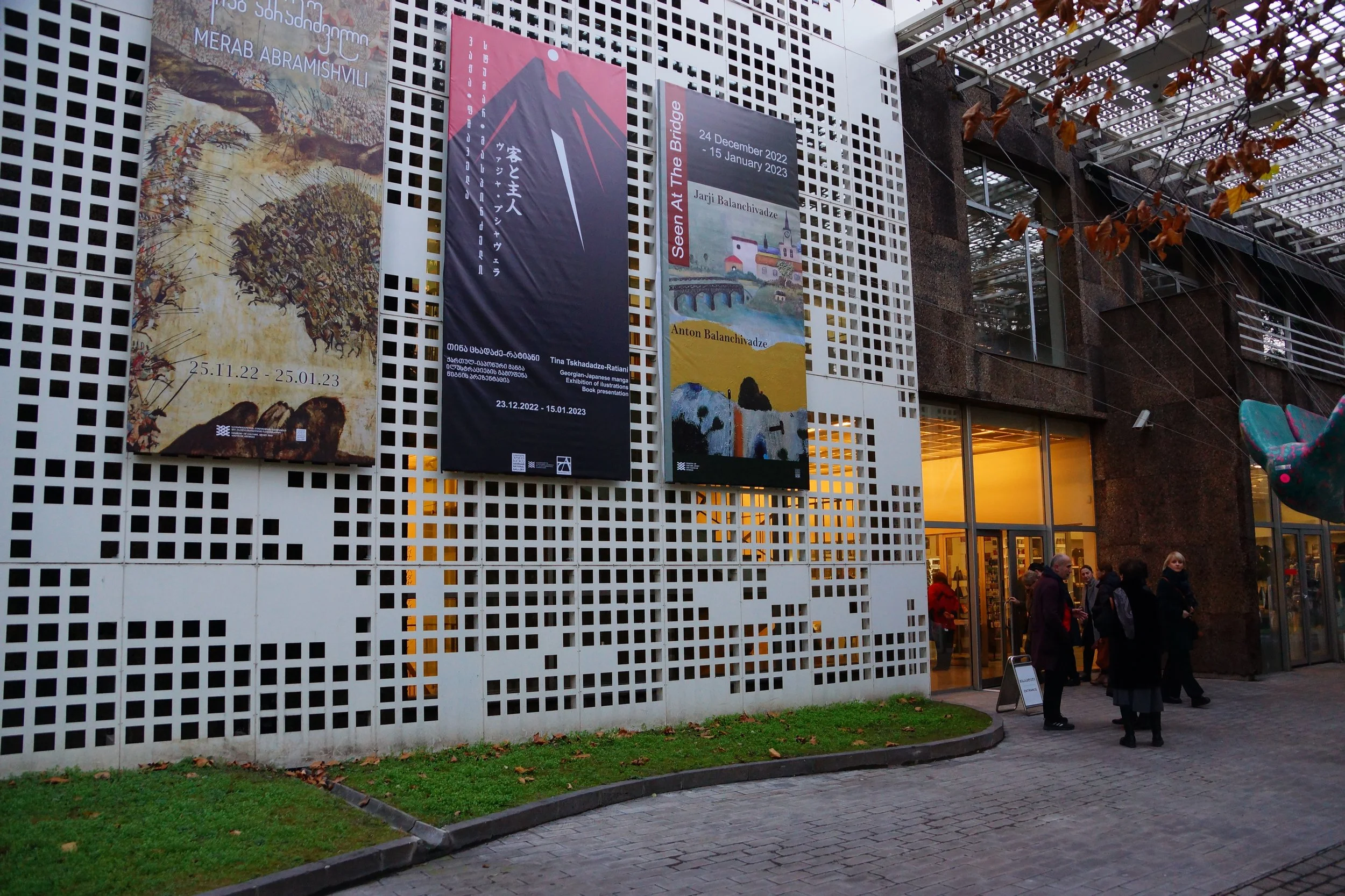 Visitors entering a georgian modern art gallery and a national museum with banners advertising art exhibitions of Jarji and Anton Balanchivadze. Glass doors reveal an illuminated interior, and some people are standing outside near the entrance.