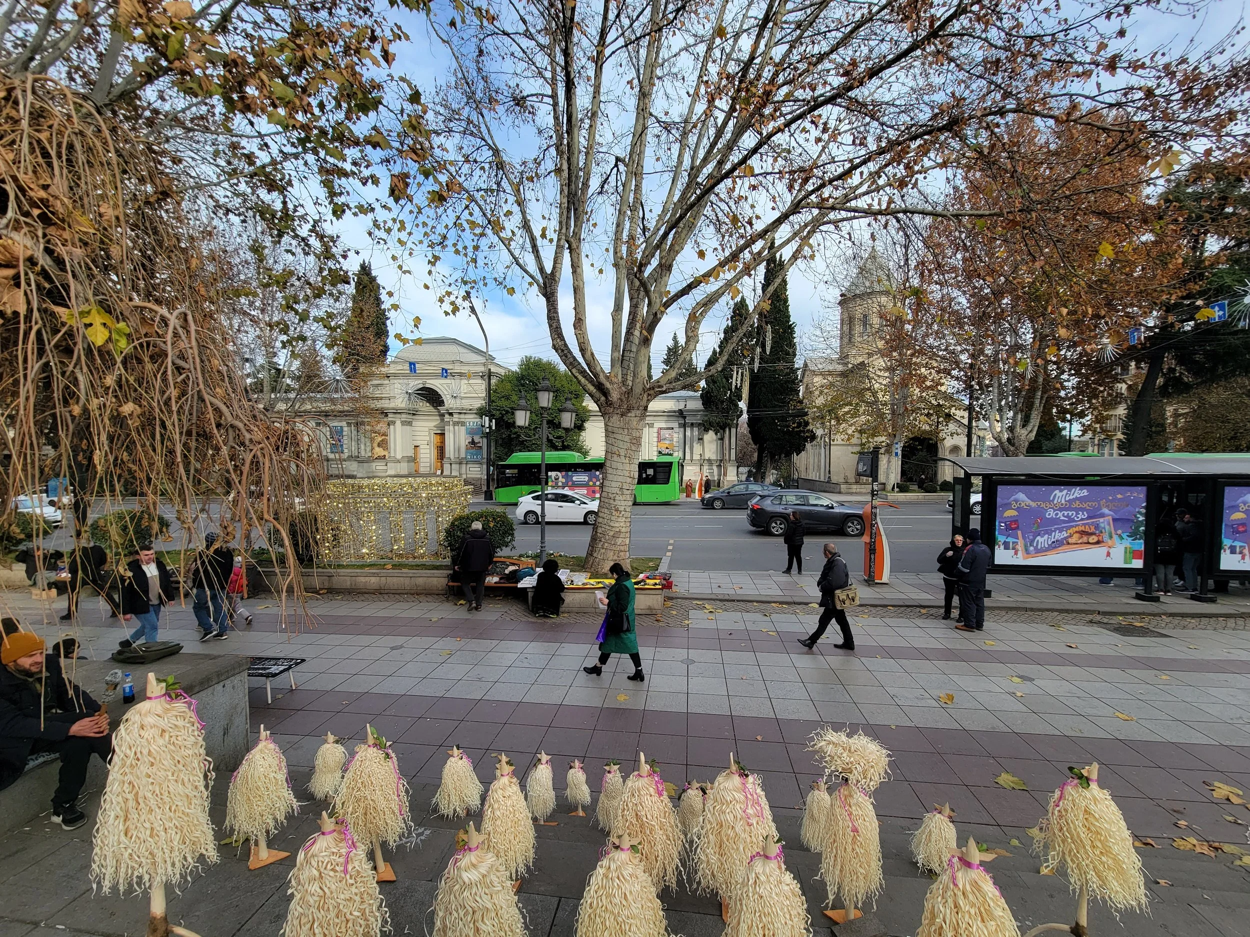 Outdoor street scene with people walking, trees with autumn leaves, a bus stop with advertisements, and handmade decorative objects on the sidewalk in front.