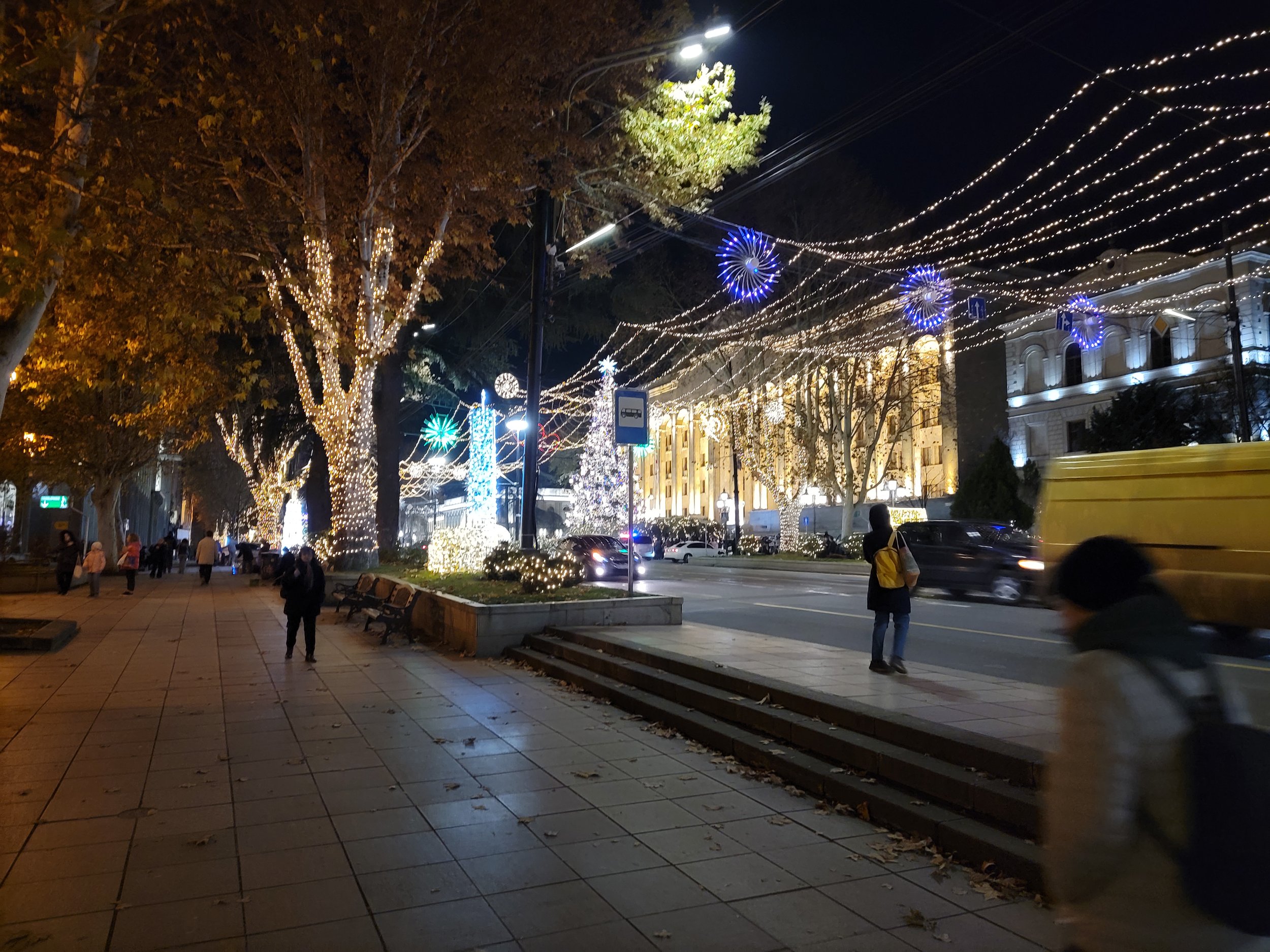 Nighttime city street decorated with Christmas lights, including trees wrapped in lights, hanging string lights, and illuminated ornaments. Pedestrians walk along the sidewalk, some with backpacks, and cars drive on the road.