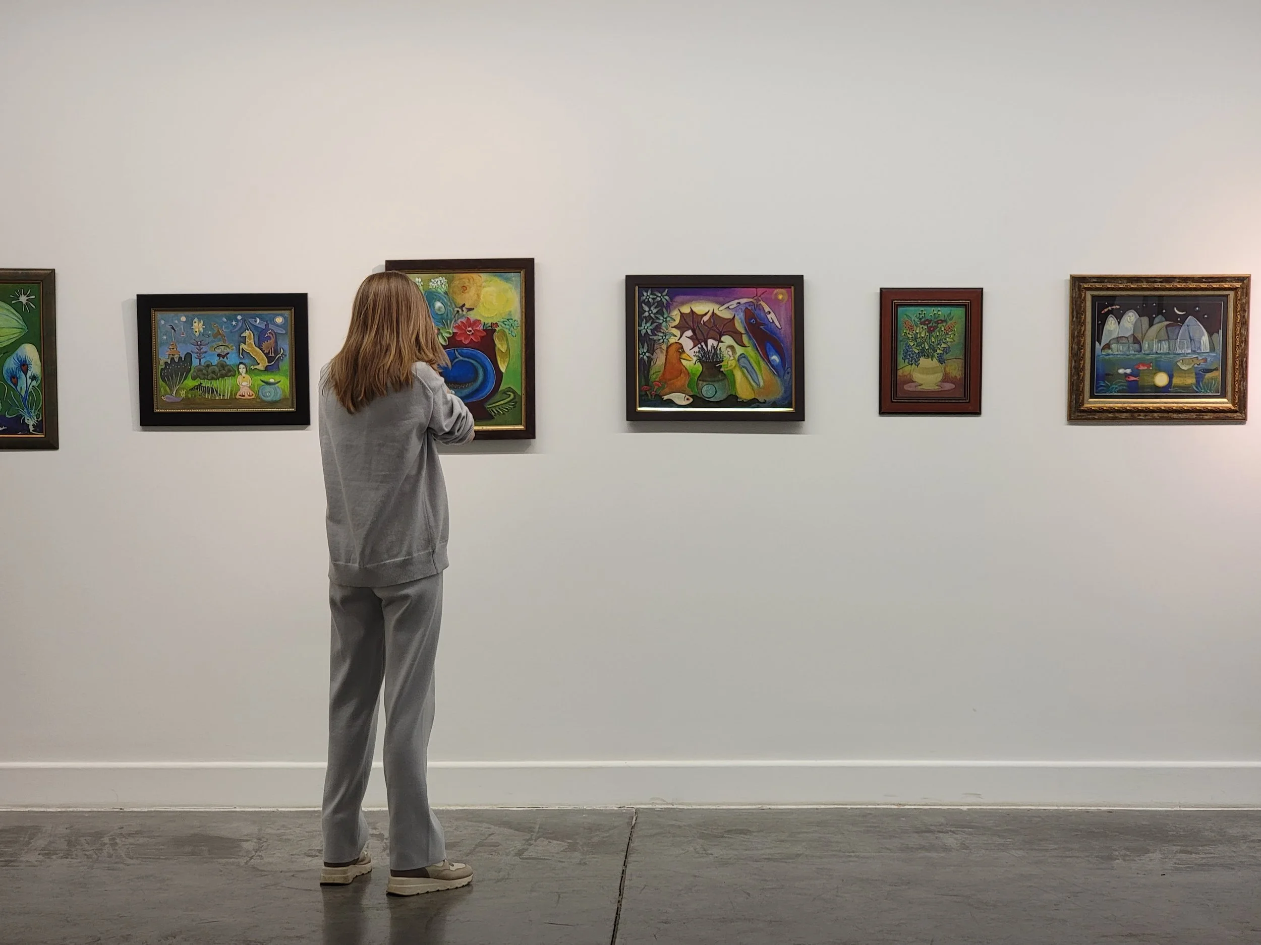 A woman with shoulder-length blonde hair wearing a light gray sweatshirt and matching sweatpants standing in front of a white gallery wall with five colorful paintings, observing one of the artworks.