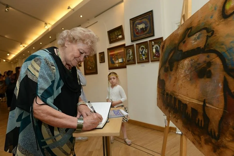 An elderly woman with curly, light-colored hair is signing a book at an art gallery. A young girl with long, blonde hair, wearing a white top and striped skirt, stands nearby. The gallery walls are adorned with numerous framed paintings, and a large 