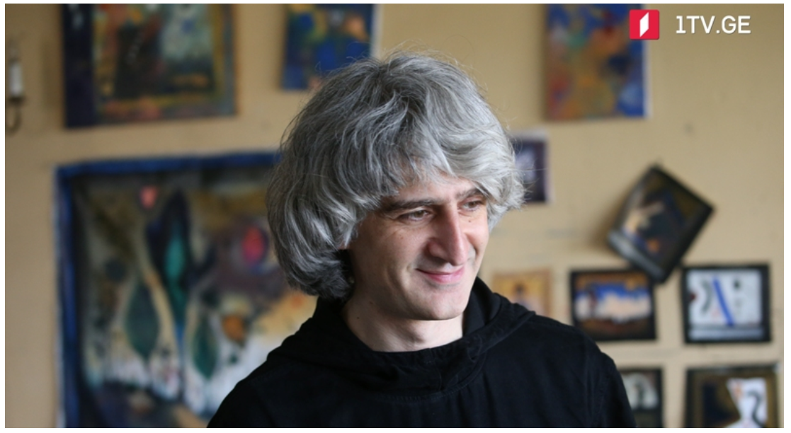 Anton Balanchivadze smiling, standing in front of a wall decorated with colorful paintings and photographs.