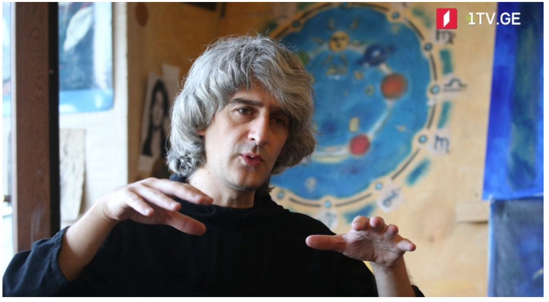A man with gray, curly hair speaking, gesturing with his hands, in front of a blue and orange abstract artwork that resembles a celestial or planetary scene, with a window and posters in the background. Anton Balanchivadze