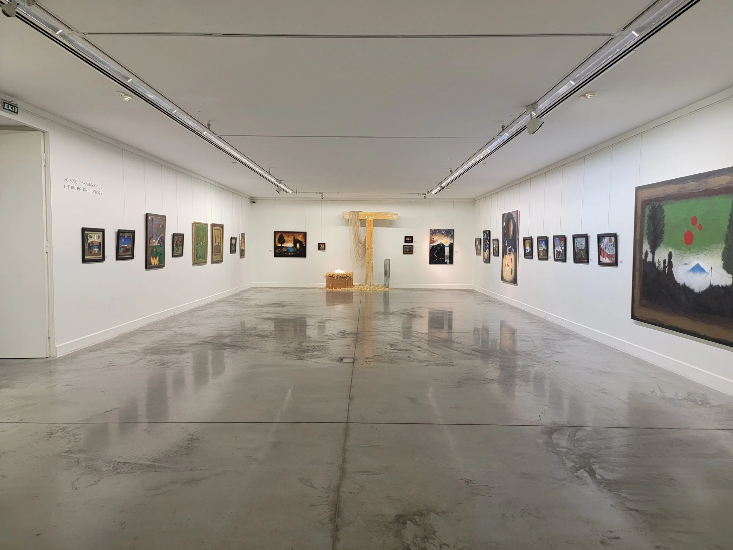 An art gallery with white walls displaying various framed paintings. At the center back, there is a large dried plant installation resembling a cross with a small bench below it. The gallery space has a polished concrete floor and ceiling-mounted tra
