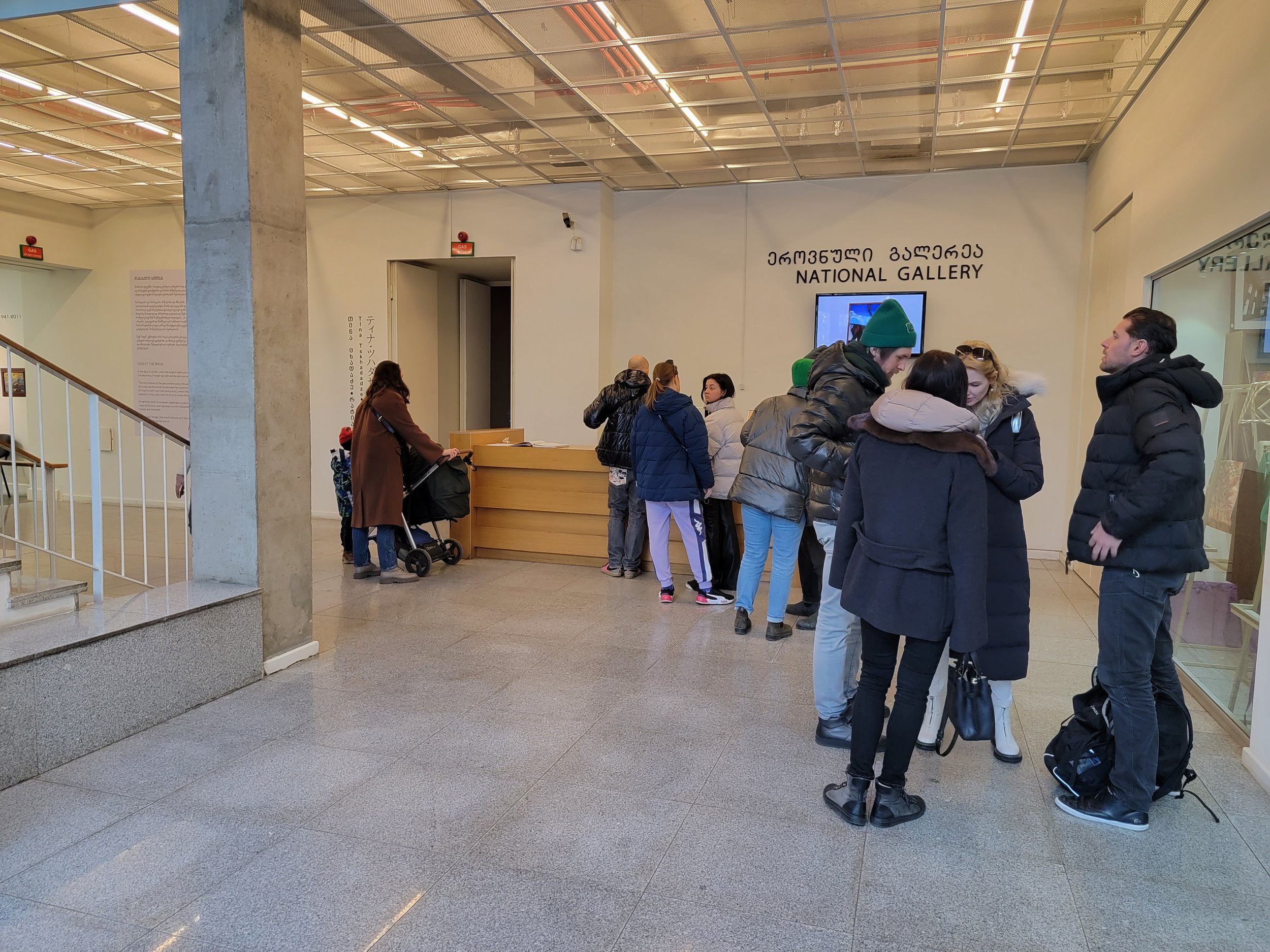 People standing in line at the ticket counter in the National Gallery with a TV screen and signage in multiple languages.