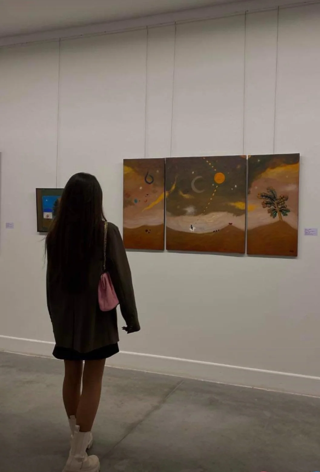 A woman with long dark hair, wearing a dark jacket, white shoes, and carrying a pink bag, is looking at a triptych painting in an art gallery. The painting depicts a surreal landscape with celestial elements like moons and stars, a tree on the right 
