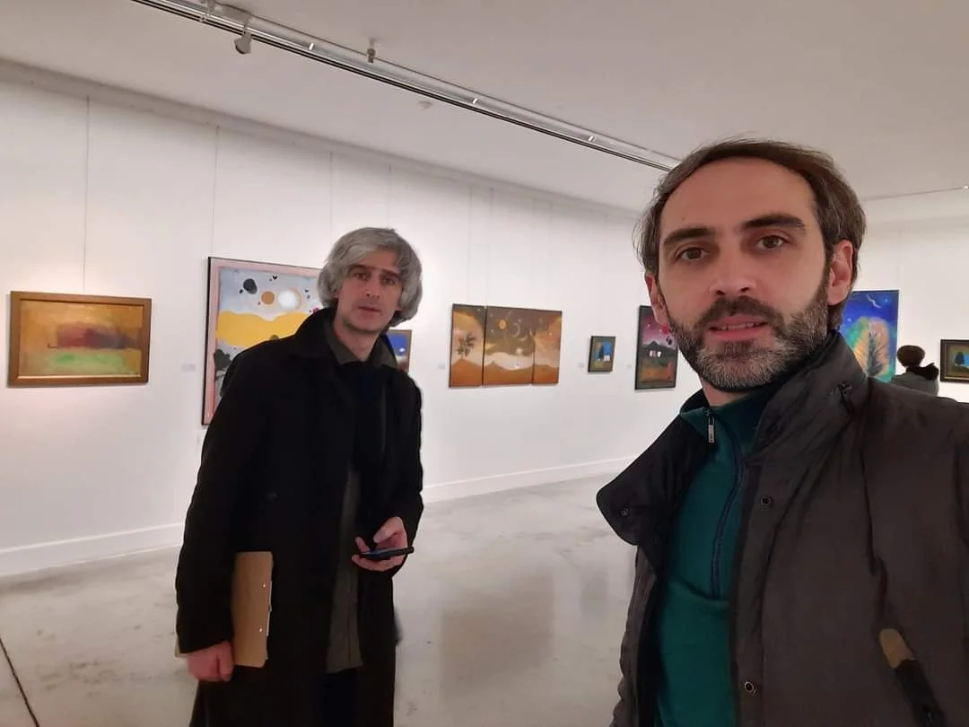 Anton Balanchivadze and Giorgi Kukhalashvili in an national gallery at the exhibition "Seen at the bridge", with colorful paintings hanging on white walls behind them.
