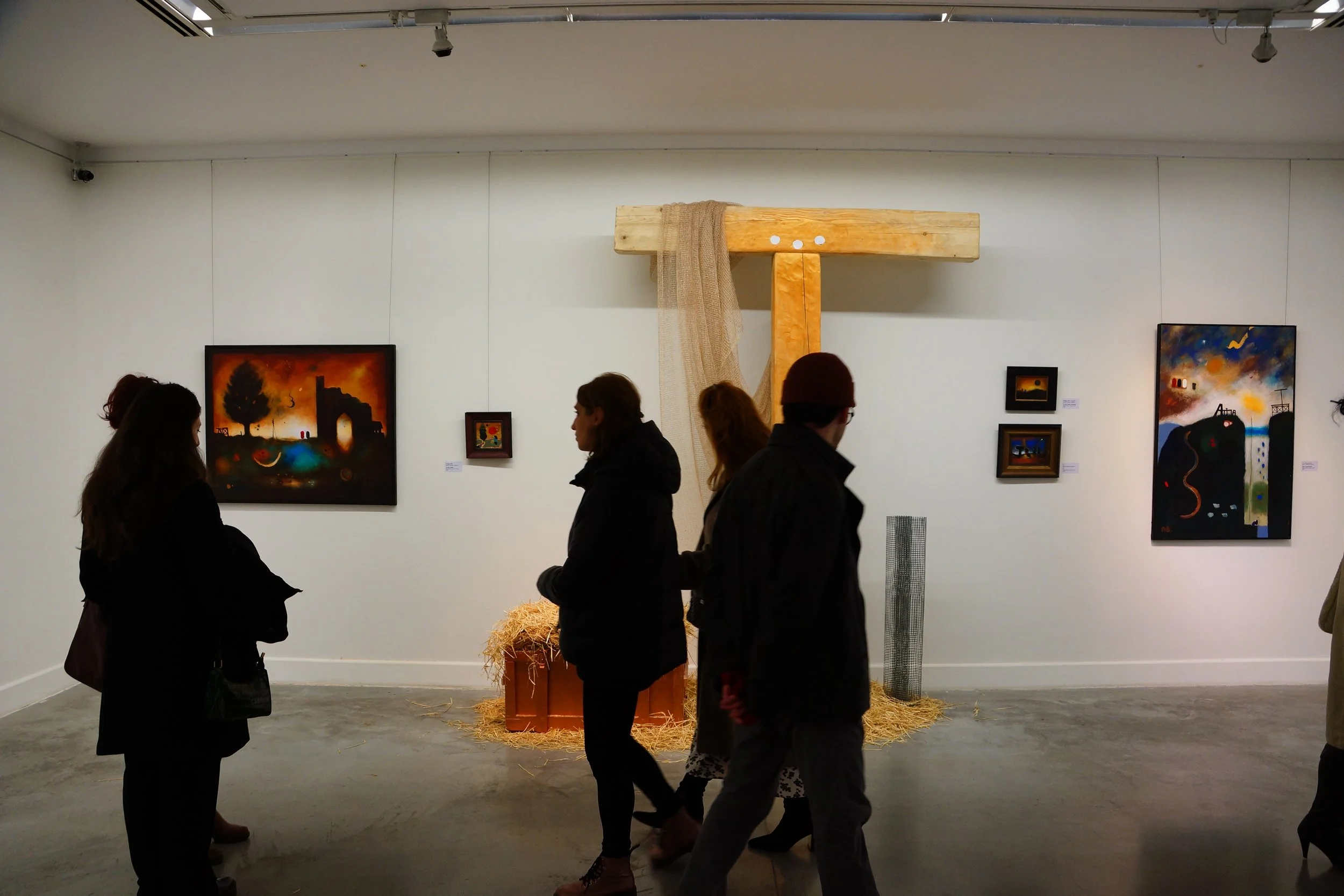 People viewing artwork in an art gallery with four paintings on the wall and wooden and straw sculpture in the center.