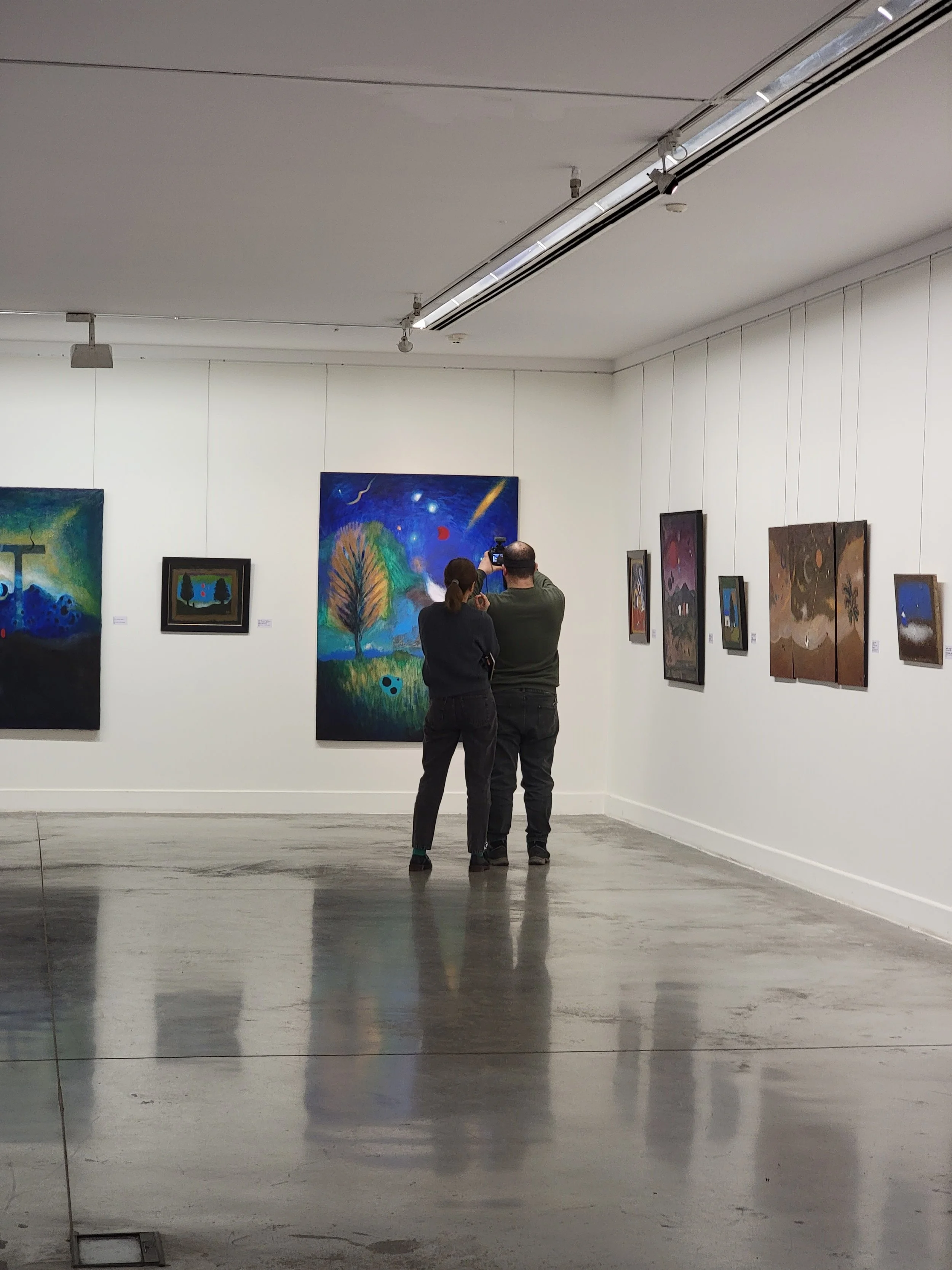 Two people stand in front of paintings of Anton Balanchivadze in art gallery, taking a photo of a colorful painting featuring a tree, a blue sky, and cosmic elements.