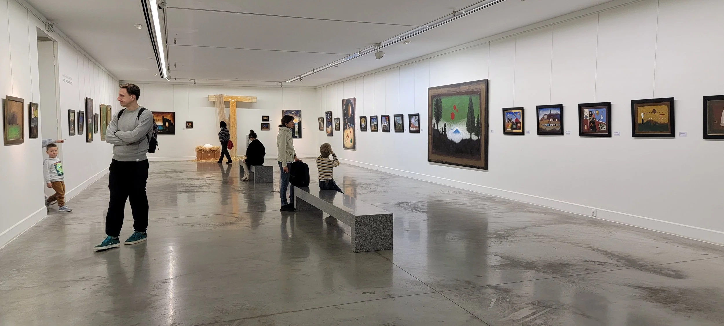 People viewing artwork in a modern art gallery with white walls and polished concrete floors. Several small framed paintings are hung along the walls, and a large, colorful painting is prominently displayed. Some visitors are seated on benches, while