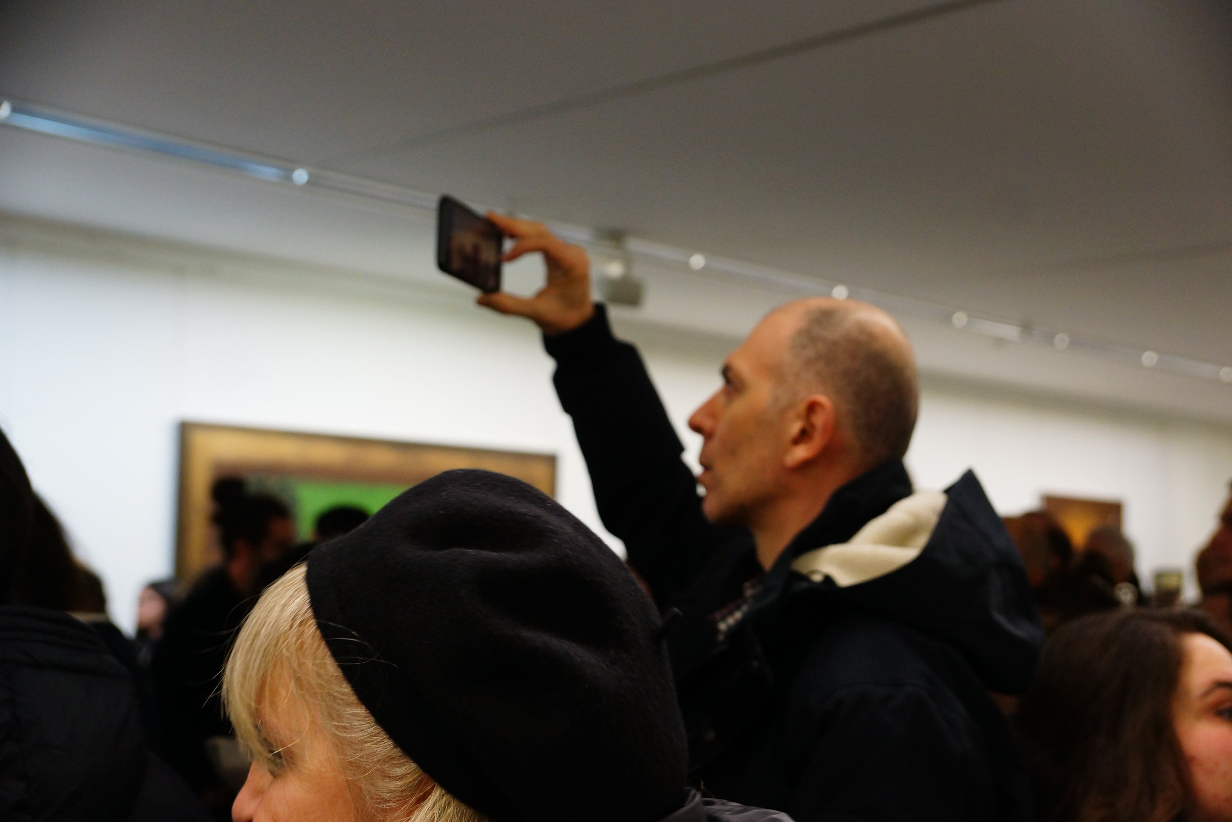 A man with a shaved head taking a photo or video with a smartphone at an indoor event, surrounded by other people with a painting on the wall behind him.