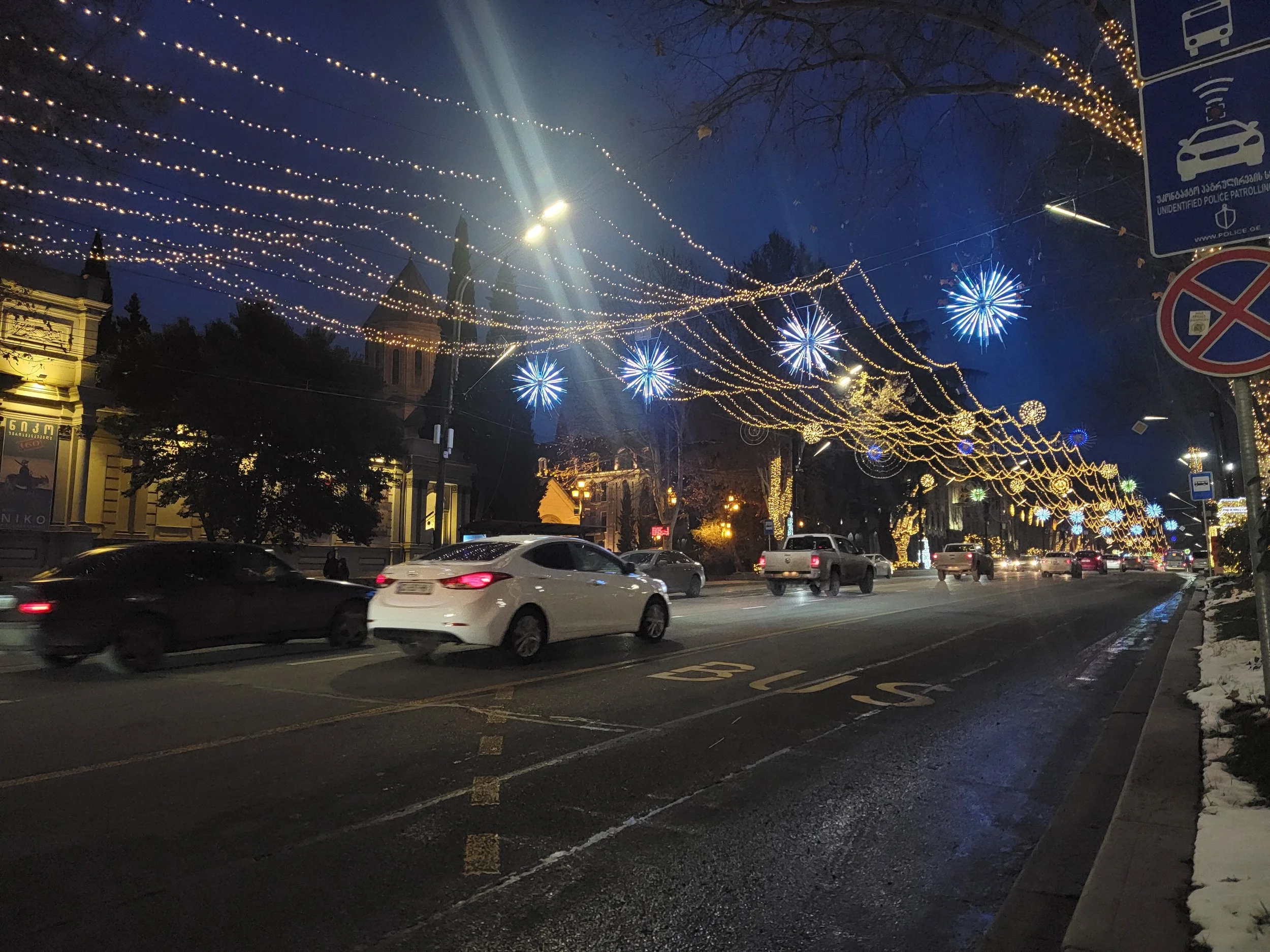 Street decorated with holiday lights and blue firework-style illuminations, with cars driving through at night.