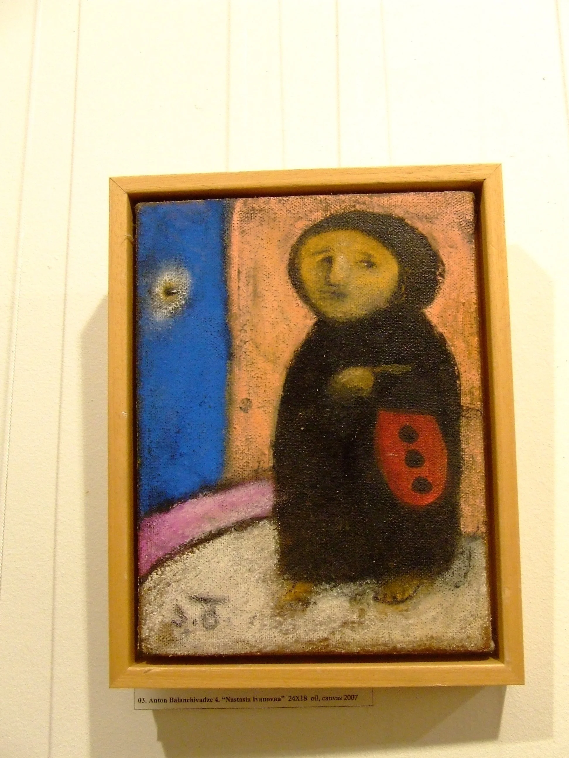 A painting of a figure with a yellow face and black body, holding a red shield with three black dots, standing on a white ground against a background of pink and blue with a small white and yellow object in the blue area.