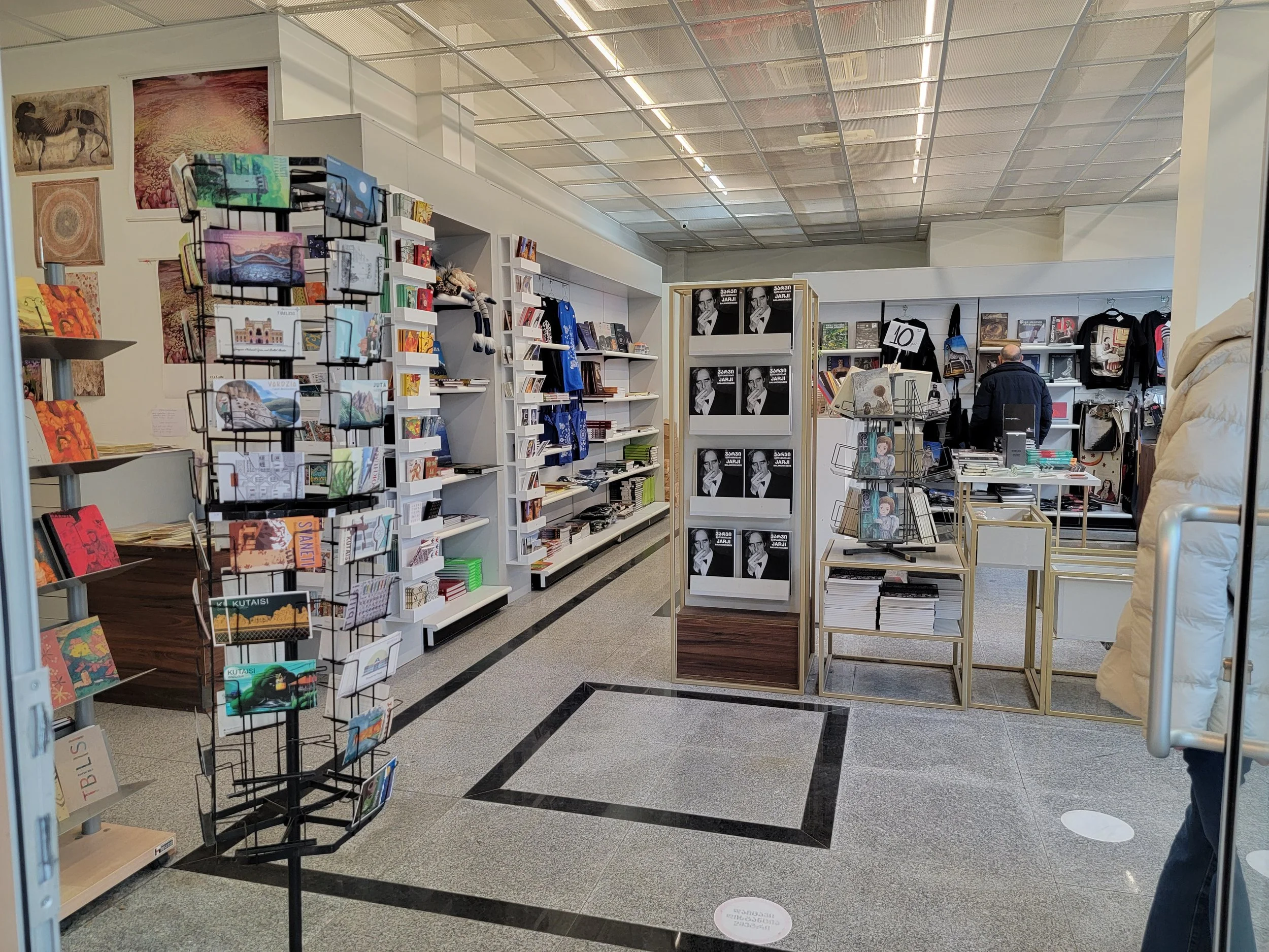 View of a bookstore aisle with various books, magazines, and merchandise on white shelves and display racks, with shoppers browsing in the background.