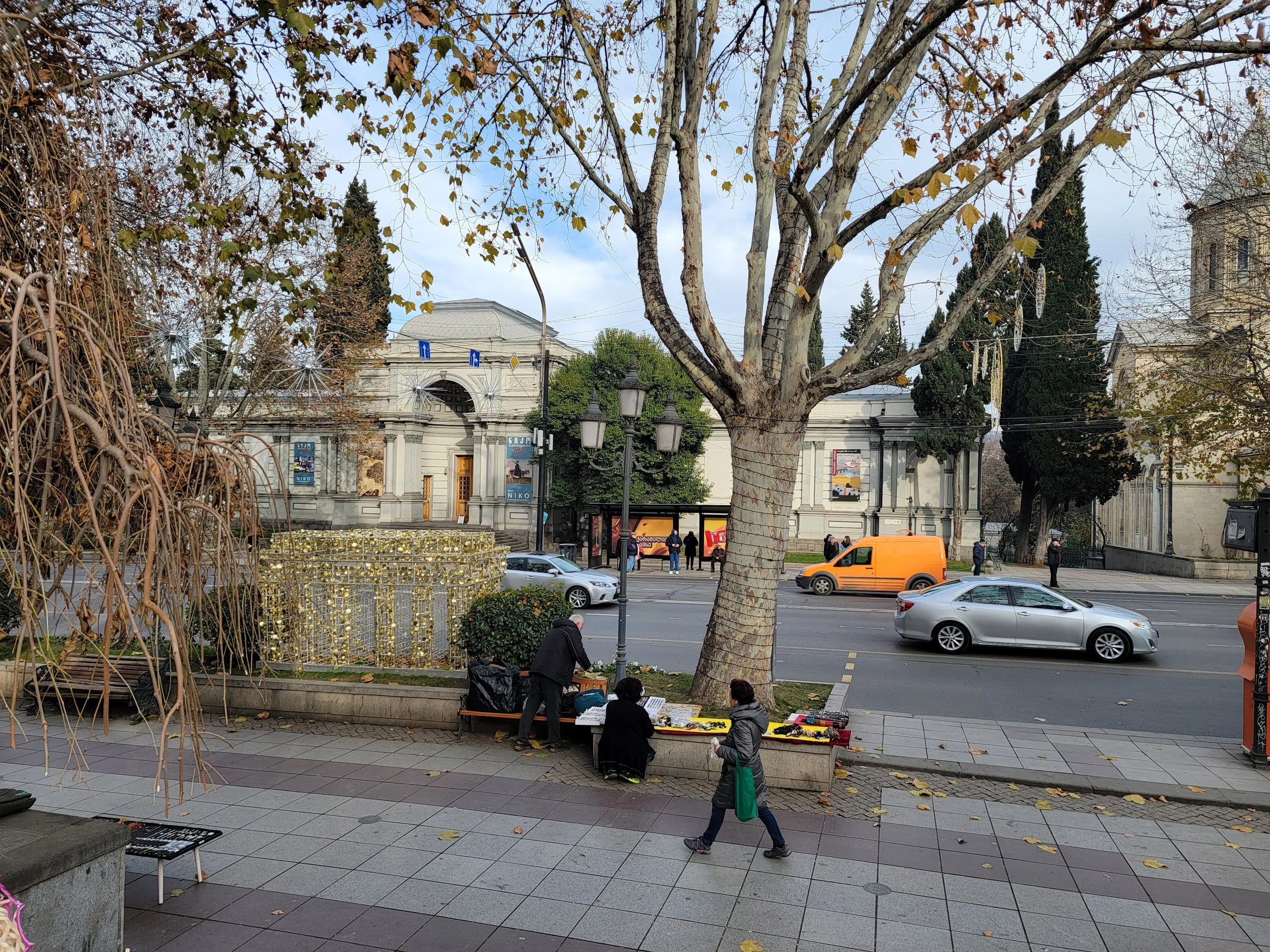 Street scene with a large tree, sidewalk vendors, parked and moving cars, and classical-style buildings in the background.