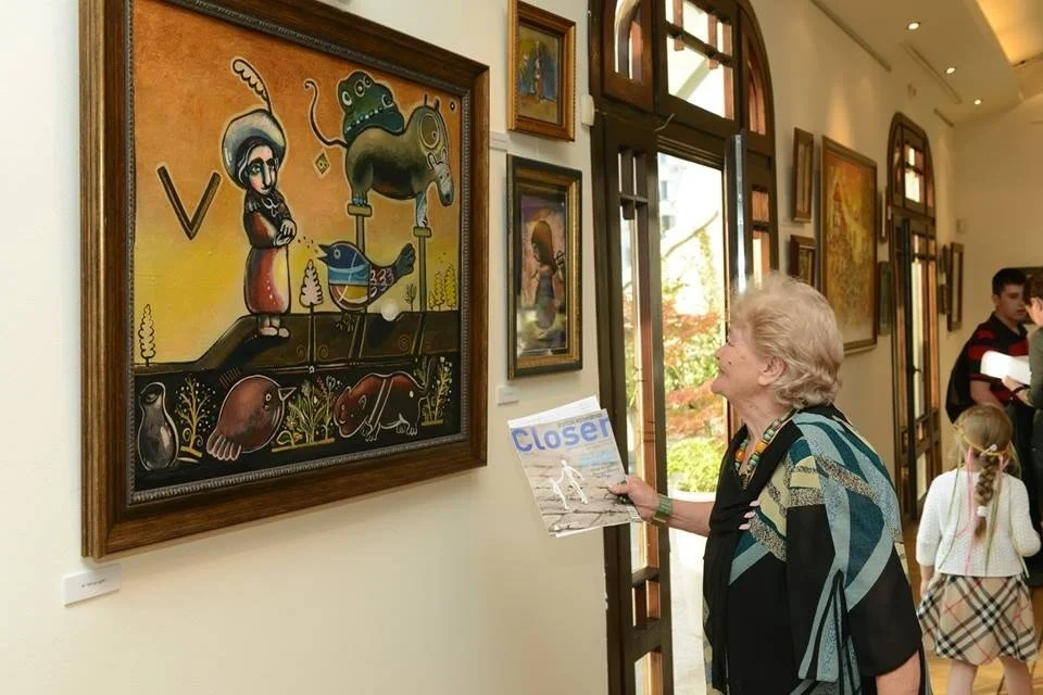 An elderly woman holding a magazine titled 'Closer' looking at a colorful painting in an art gallery. The painting features a stylized figure with an umbrella, a fish, and various abstract elements. There are other framed artworks on the wall, and la