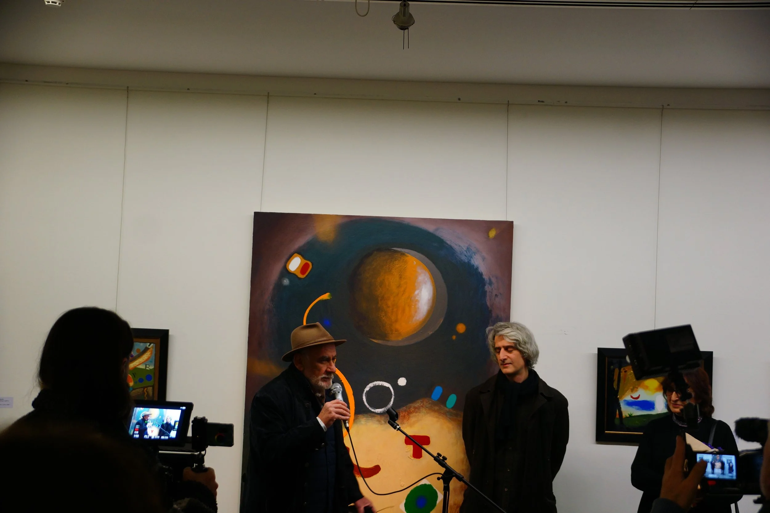 Two men are speaking at an art gallery with paintings on the wall behind them. The man on the left is wearing a fedora and speaking into a microphone, while the man on the right has gray hair and is listening. Photographers are capturing the moment.