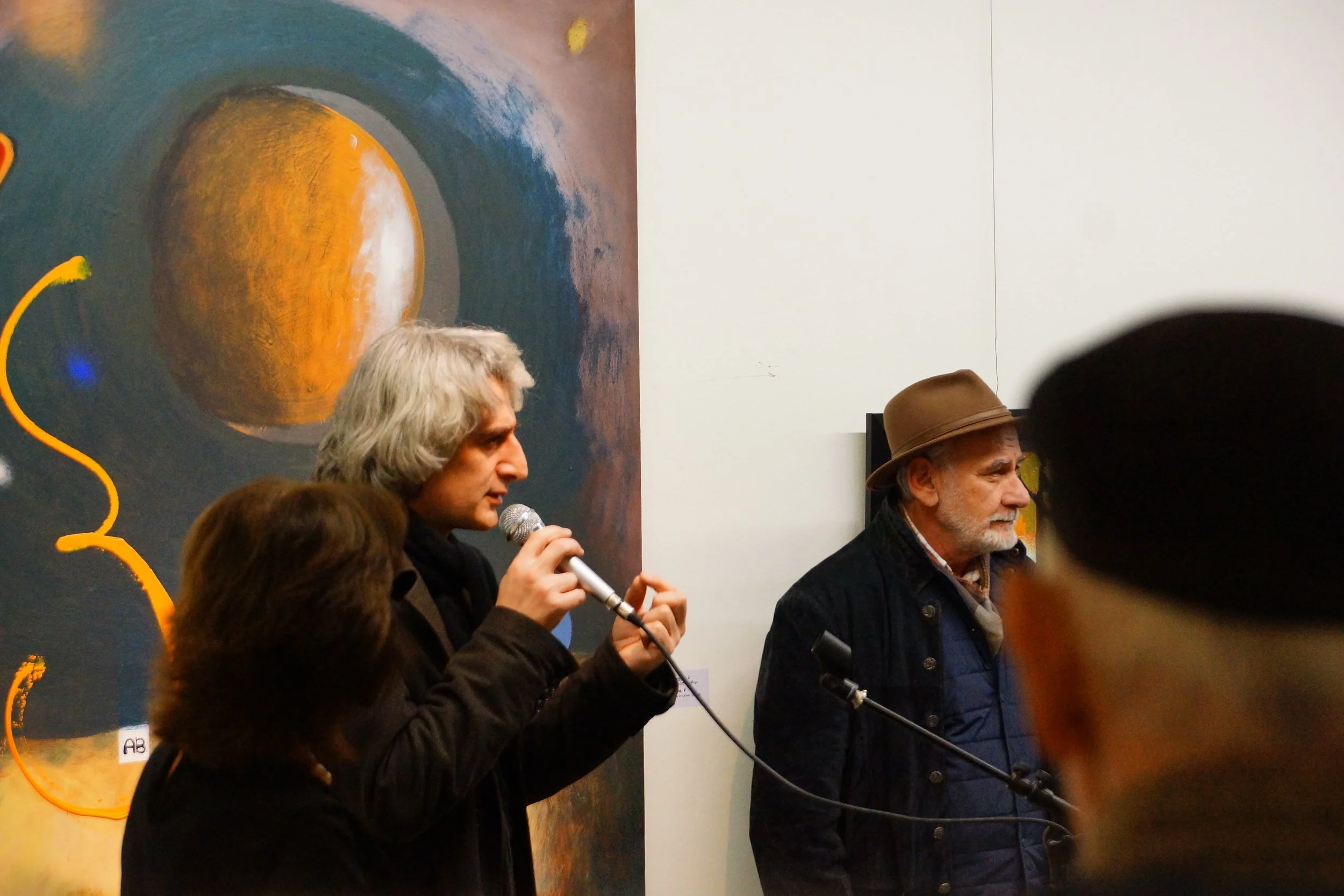 A man with gray hair speaking into a microphone at an art exhibition, with a large abstract painting featuring a circular shape in orange and brown hues behind him, and another man wearing a brown hat and dark jacket standing nearby.