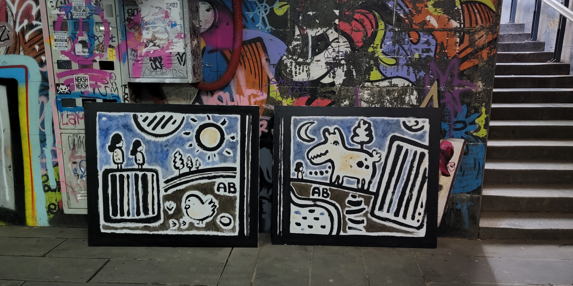 Street graffiti art on two panels featuring cartoonish drawings of a girl and a duck on the left panel, and a dog on the right panel, with a colorful graffiti-covered wall and staircase in the background.