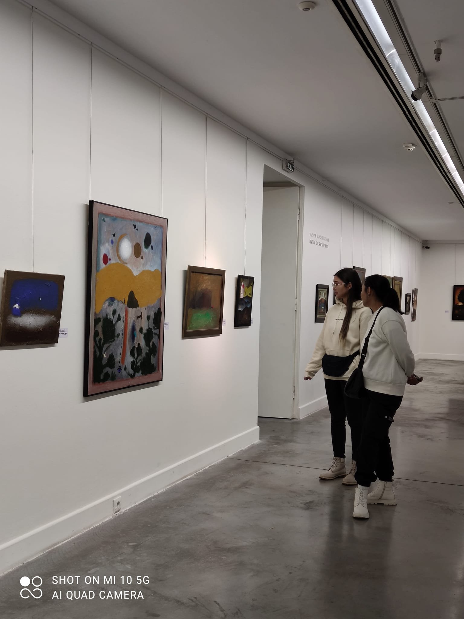 Two women are looking at paintings in an art gallery with white walls and a polished concrete floor.