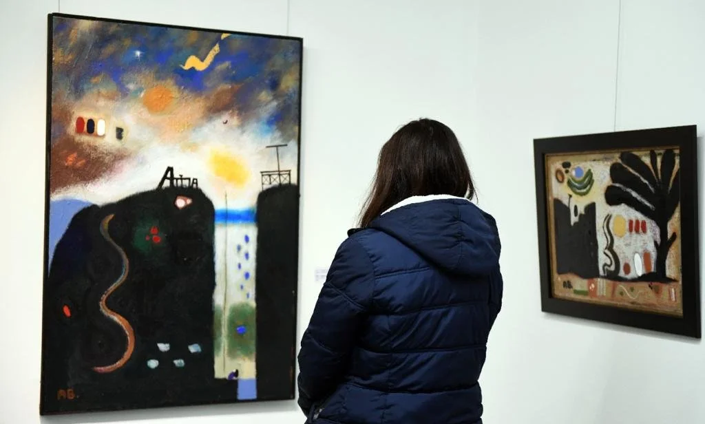 A woman with dark hair wearing a blue jacket looking at abstract paintings in a gallery. The left painting features dark and colorful abstract shapes with a cloudy sky background, while the right painting has a more structured abstract design with black, green, yellow, and red accents.