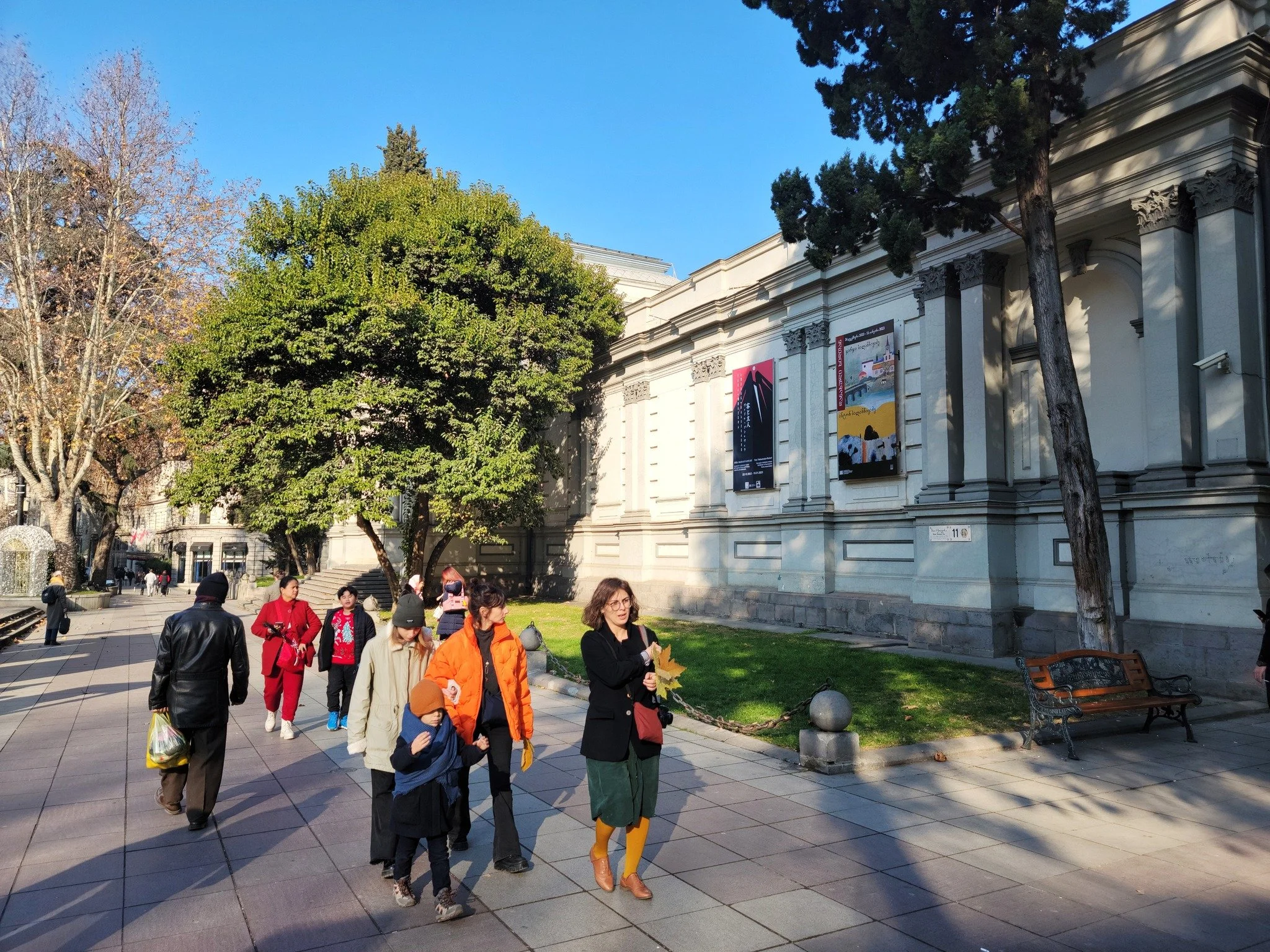 People walking on a city sidewalk in front of a large, ornate building with posters on the wall, trees, and benches.