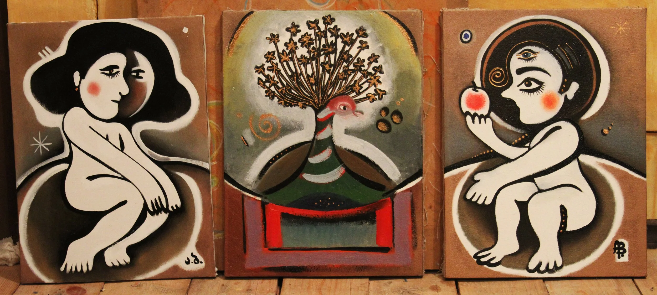 Three paintings side by side with a similar style. The left and right paintings depict cartoonish female figures with dark hair, sitting, with blush on their cheeks. The center painting features a stylized peacock with a large, elaborate tail, set ag