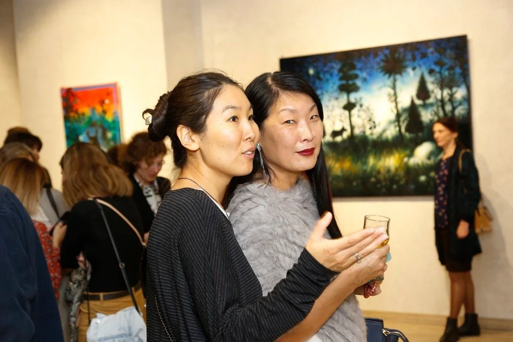 Anton Balanchivadze's Exhibition "The Gardens" at BAIA gallery , two women at an art gallery, one with dark hair in a braid and the other with long dark hair, wearing makeup and holding a drink, standing in front of colorful paintings on the wall 