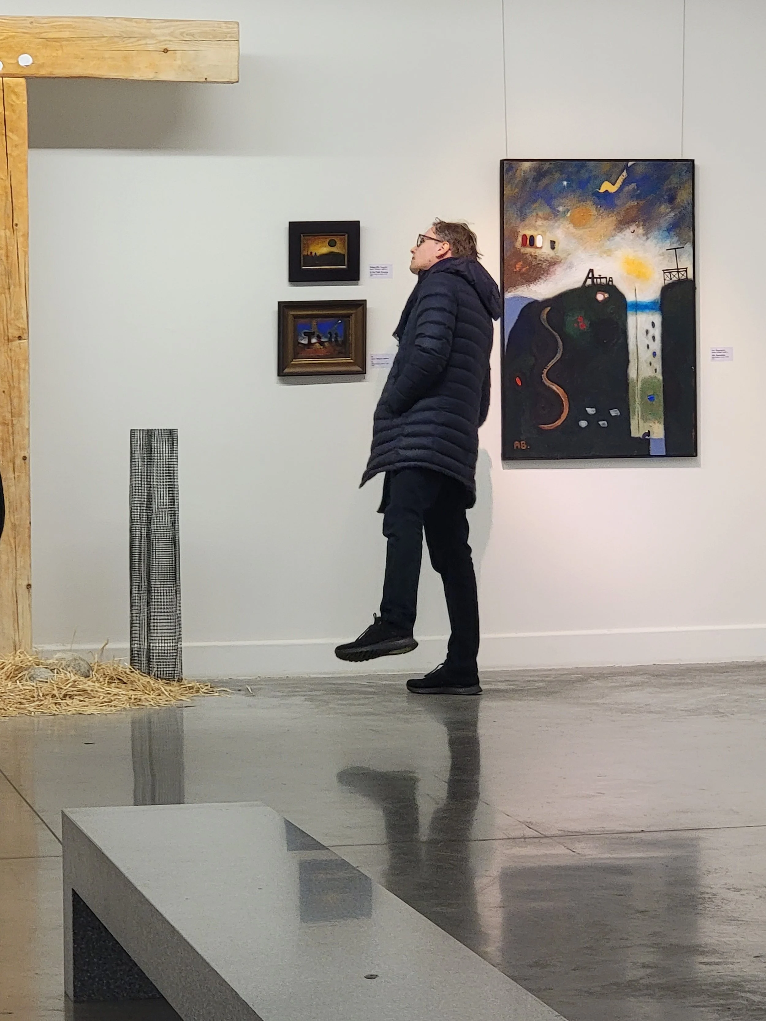 A man in a black puffer jacket and glasses is standing in an art gallery, looking at paintings on the wall.