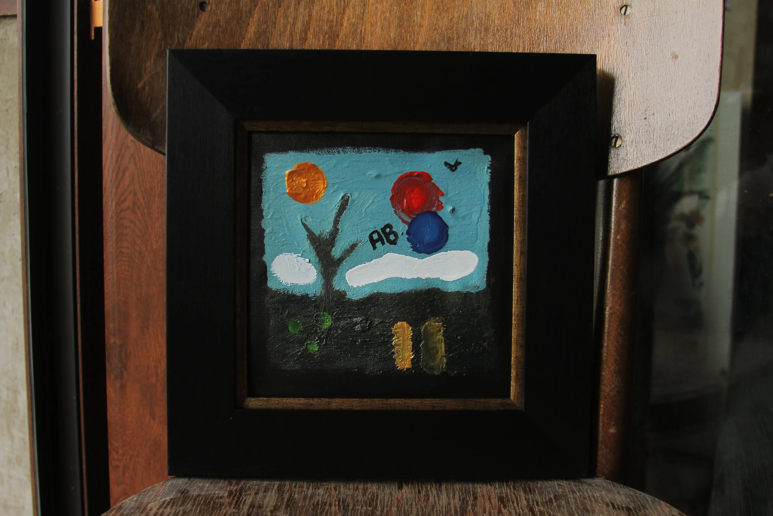 A small framed painting featuring a tree, clouds, and colorful circles on a blue background, with the text 'AB' written on it.