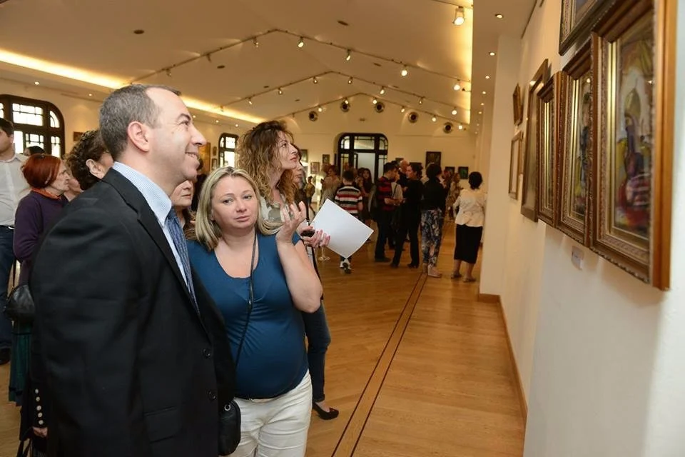 People visiting an art gallery, observing paintings on the wall, with a well-lit room and wooden floor.
