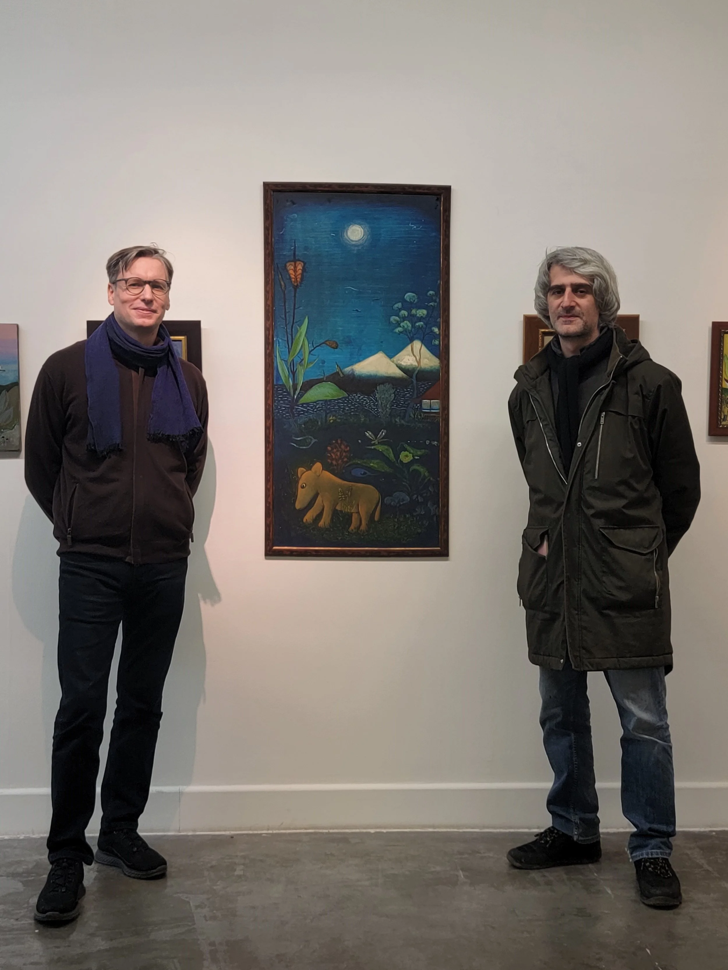 Two men standing on either side of a colorful painting of a night scene with mountains, trees, and a bear, displayed on a white gallery wall.
