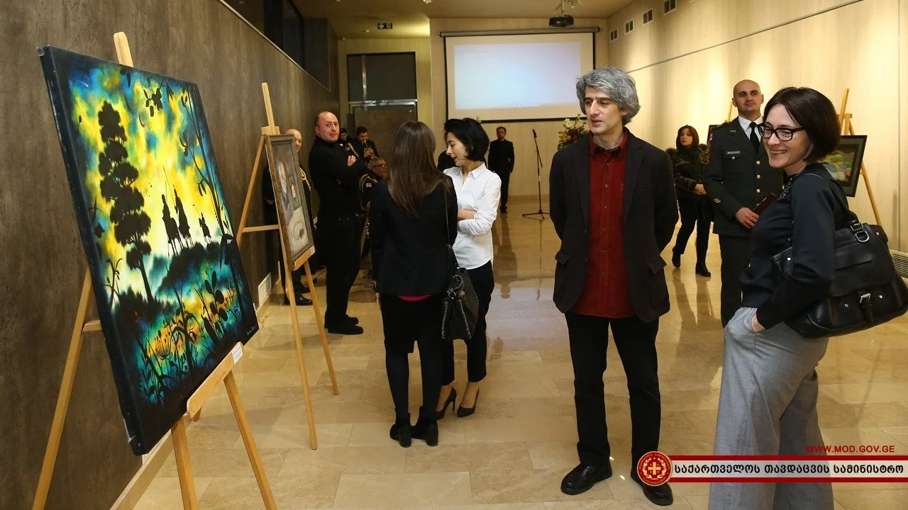 People viewing and discussing paintings displayed on easels at an art gallery or exhibition, with a projector screen in the background.