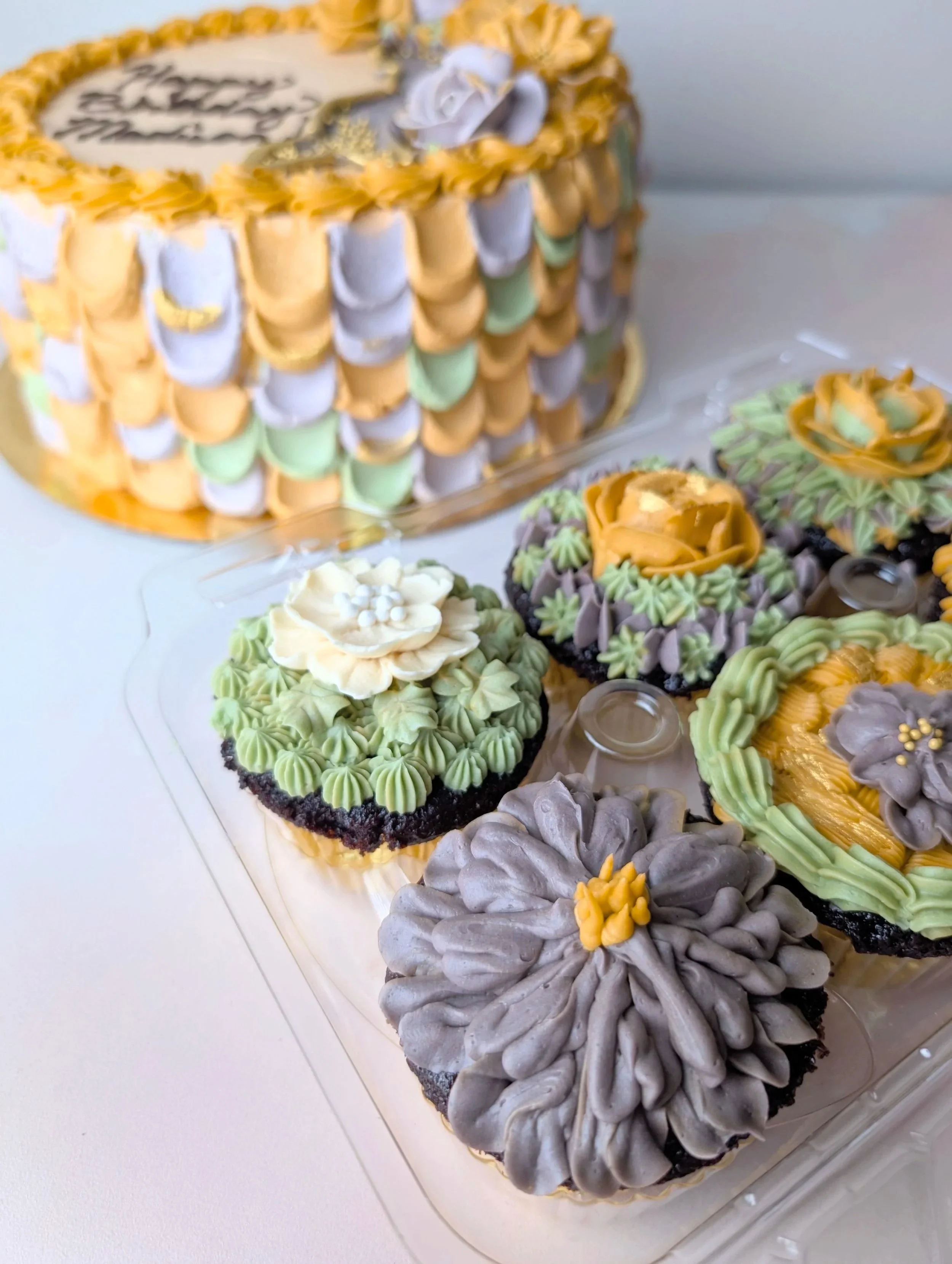 Decorative birthday cake with colorful piping, topped with a white message plaque, surrounded by cupcakes with piped icing and small flowers.