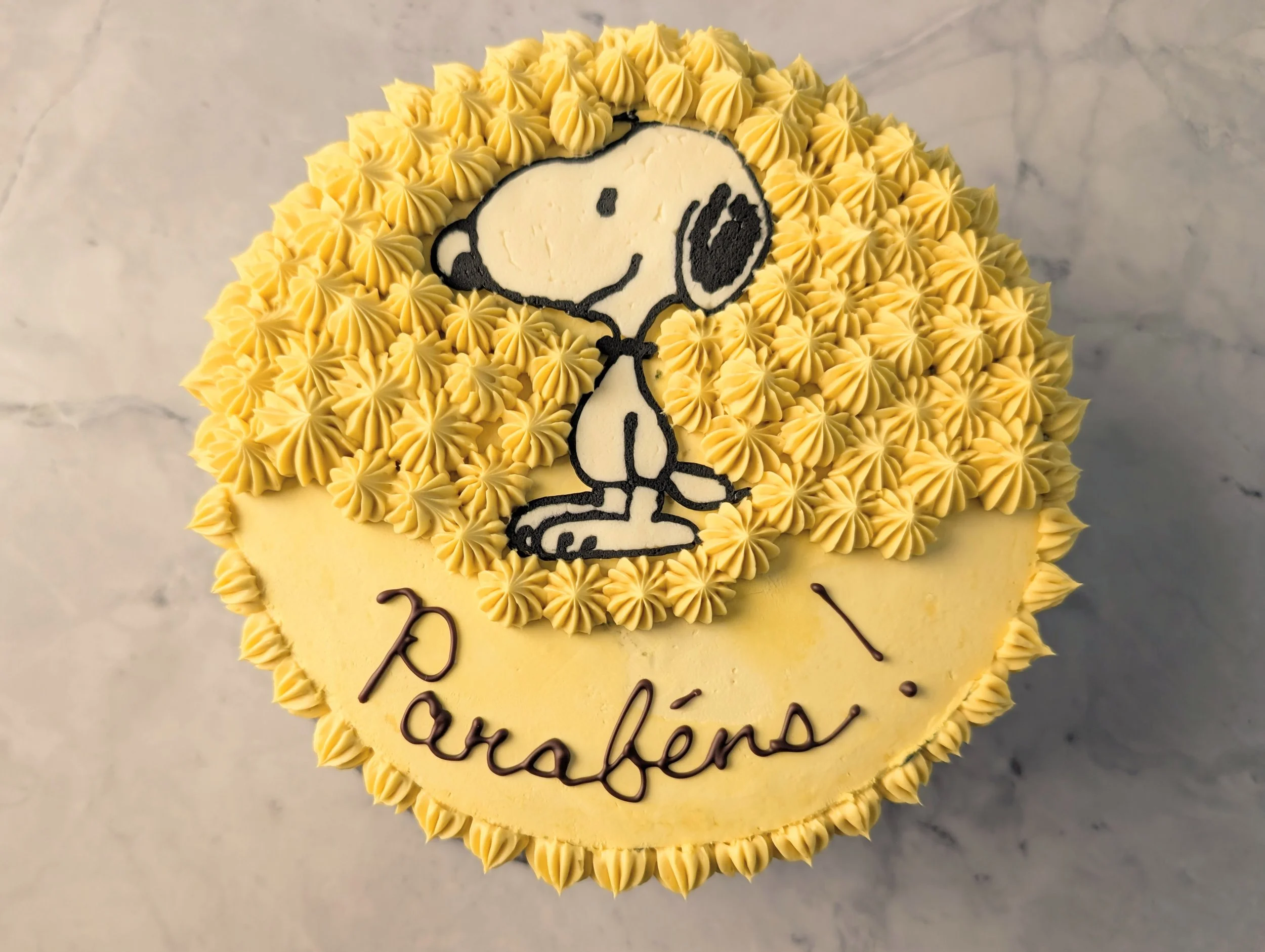 A round yellow birthday cake decorated with yellow piped frosting around the edges. The cake has an edible image of Snoopy, the cartoon dog, in the center, along with some cursive writing