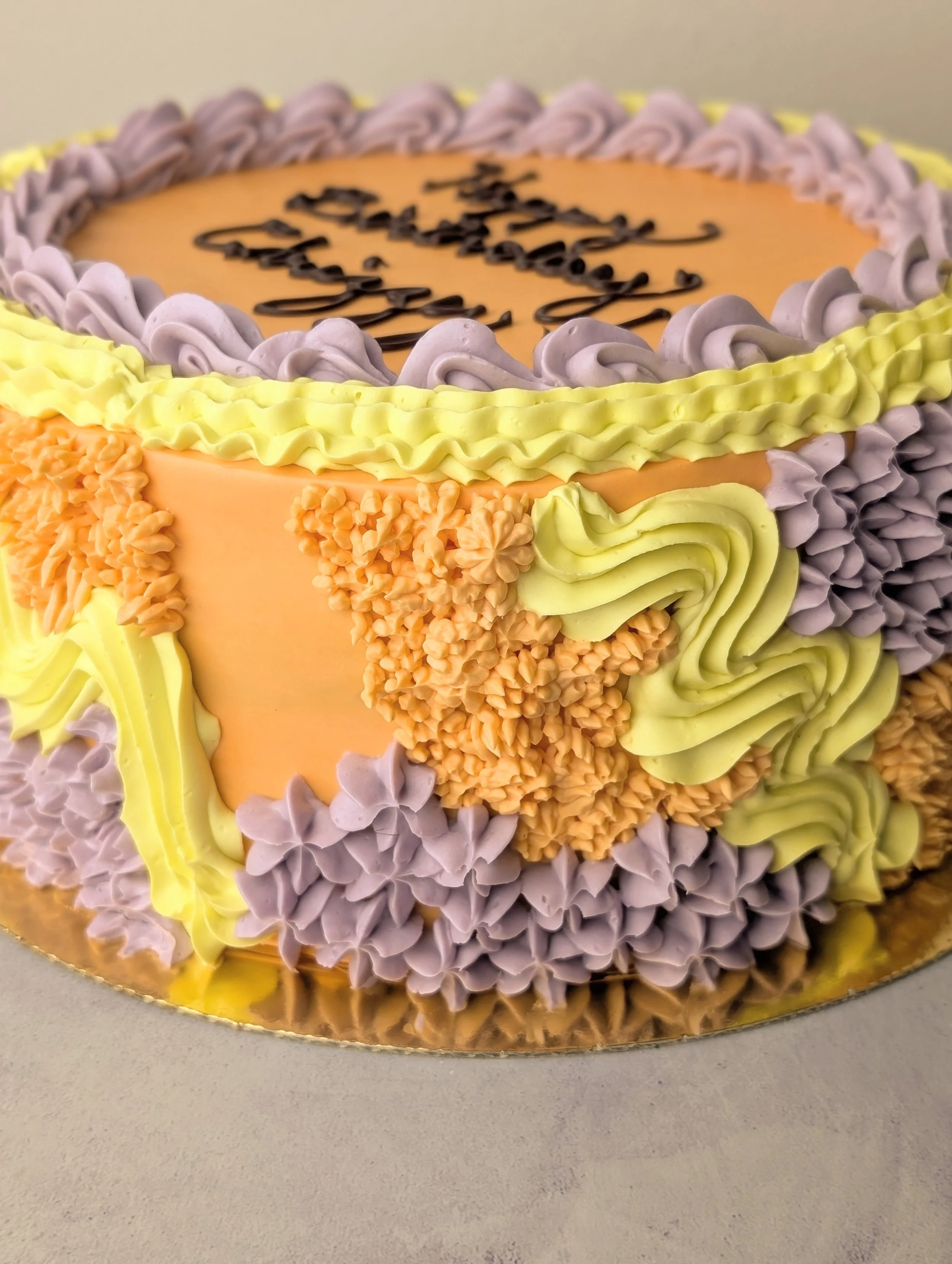 A decorated birthday cake with purple, yellow, and orange frosting, and the message 'Happy Birthday' written on top.