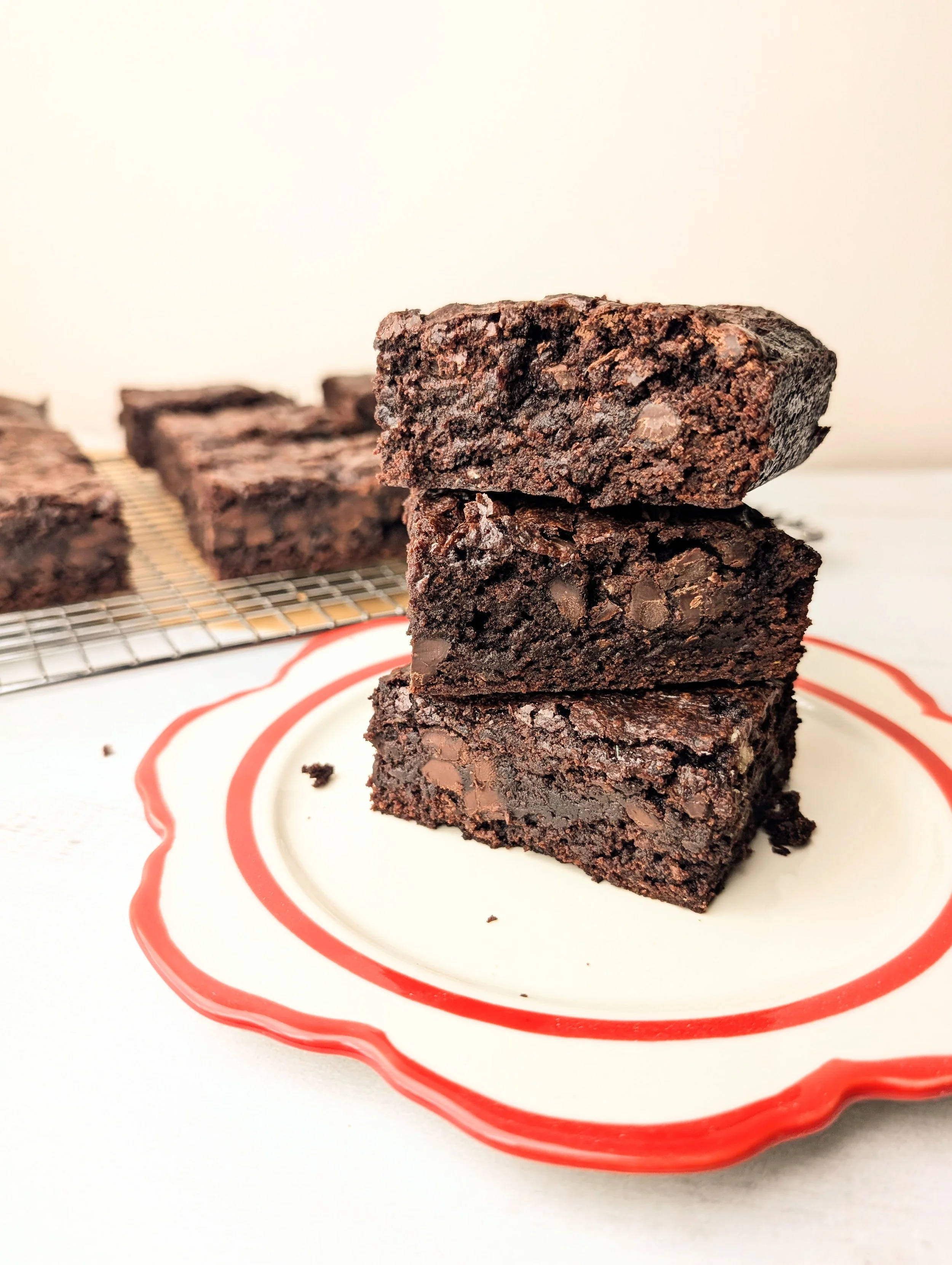 Close-up of three stacked brownies on a decorative white plate with a red rim. In the background, more brownies are cooling on a wire rack.