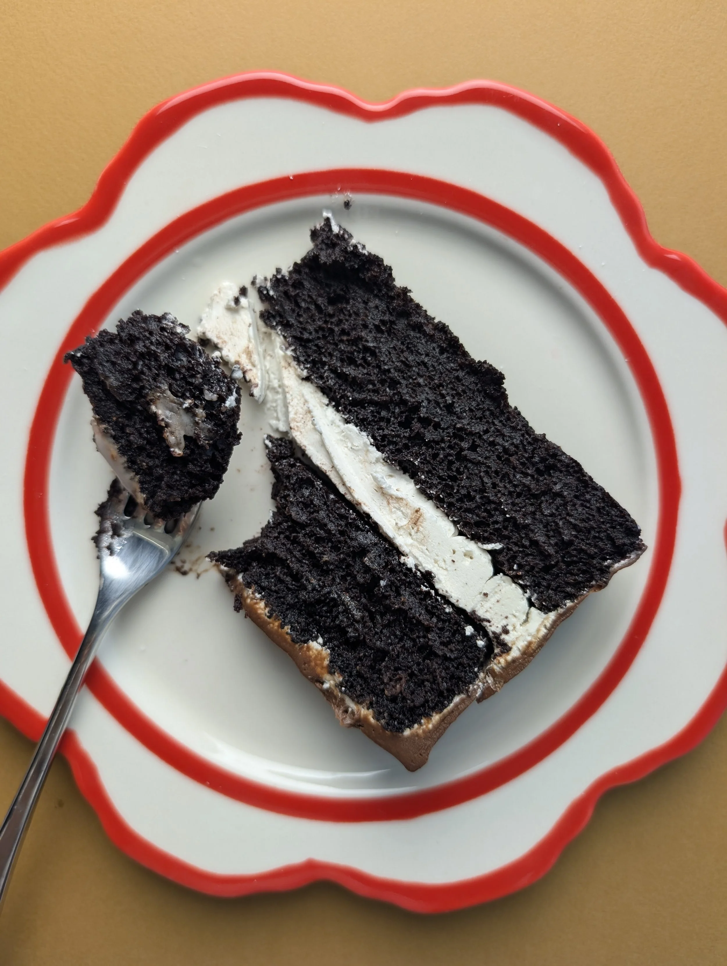 A slice of black chocolate cake with white cream filling on a white plate with a red border, with a fork resting on the plate.