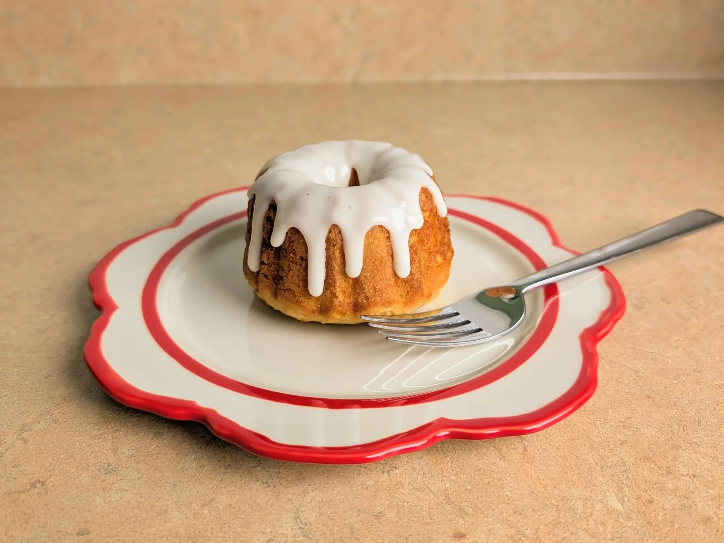 A small bundt cake with white icing on top, sitting on a decorative white plate with red edges, next to a silver fork, on a beige surface. The background is plain beige.