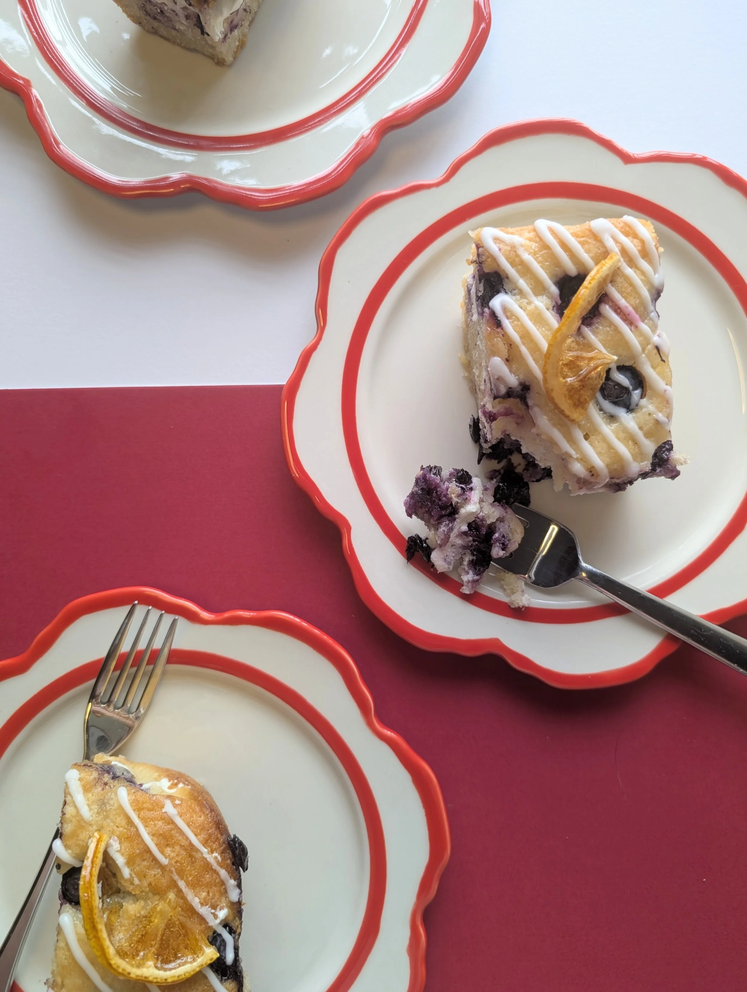A slice of blueberry dessert topped with icing and lemon slice on a red-rimmed plate, with a fork and another similar plate partially visible, placed on a pink and white surface.